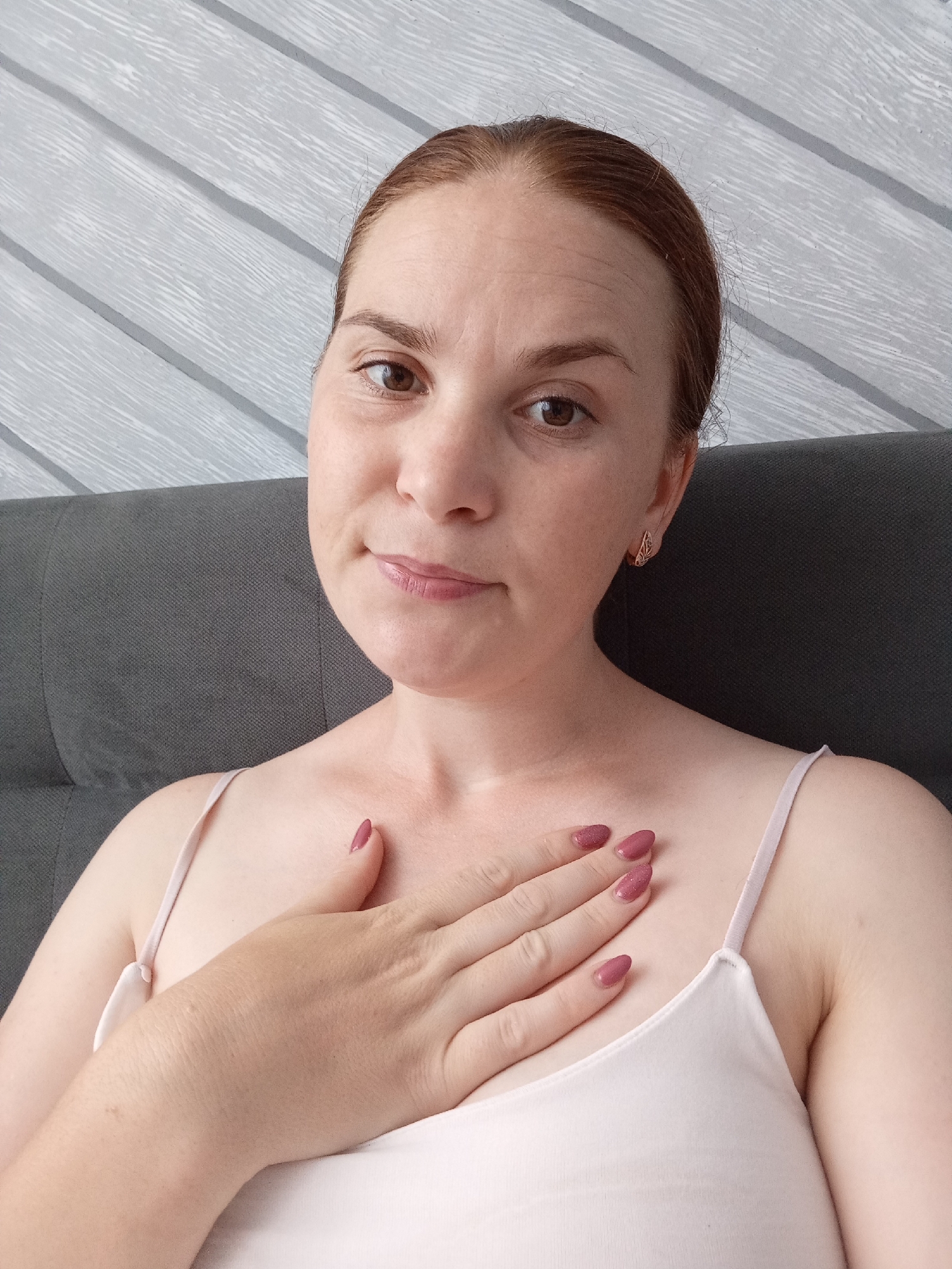 A woman with auburn hair pulled back is seated against a grey headboard, looking at the camera. Her right hand is resting on her chest, fingers spread. She is wearing a light pink camisole. The background is a grey wall with horizontal wood-paneled texture. The scene appears to be indoors, possibly in a bedroom. The specific location of Pănășești, Moldova, is not determinable from the visual information provided in the image.