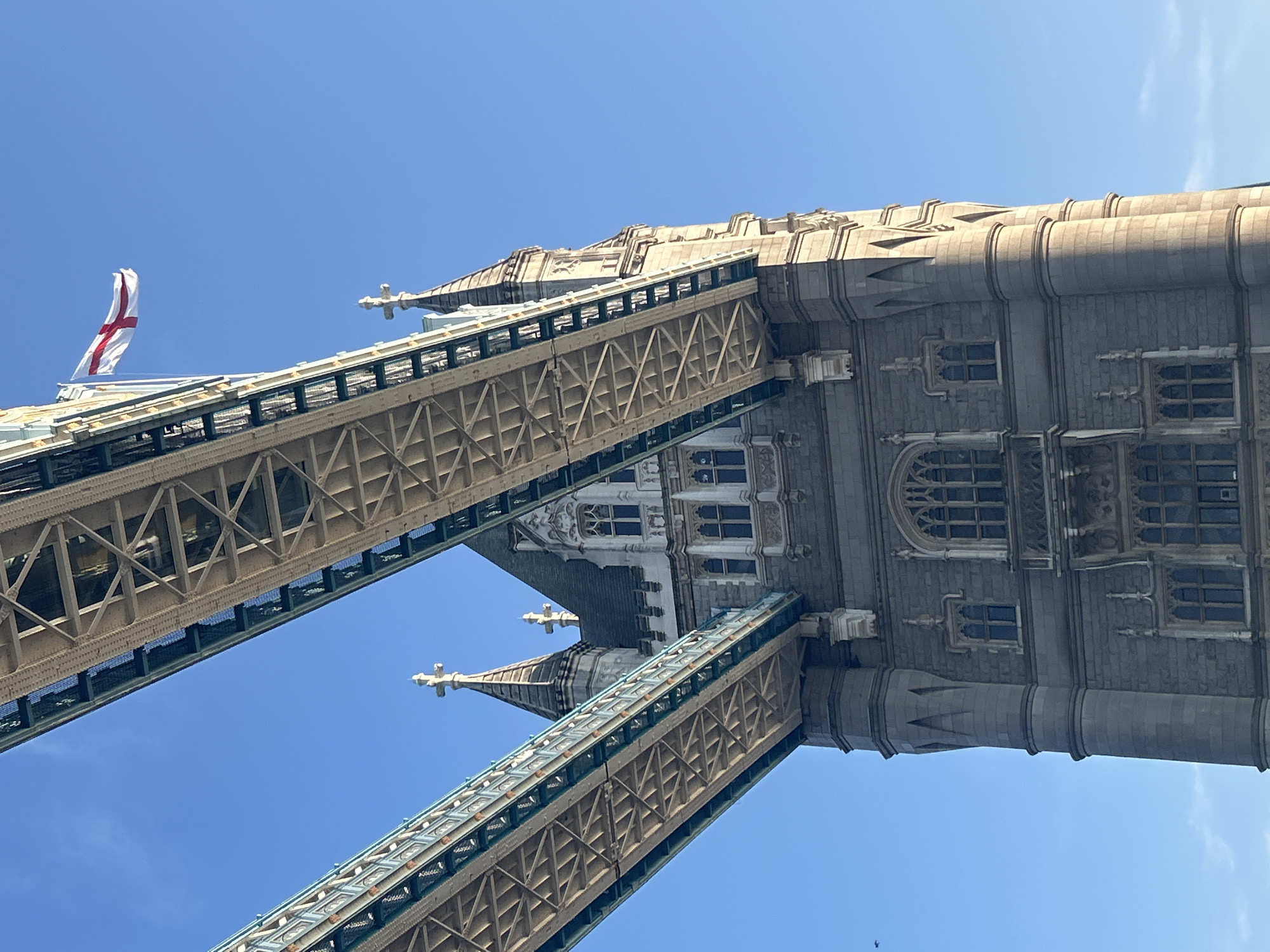This image shows a low-angle view of the Tower Bridge in London, United Kingdom, against a clear blue sky. The bridge's iconic Gothic Revival style towers and ornate stonework are visible, as are the steel structures of the bridge walkways. An English flag flutters from a flagpole on the right tower. The architectural details of the towers, including arched windows and decorative crenellations, are prominent. The lighting suggests it is either early morning or late afternoon, with the sun casting shadows on the stonework. No people are visible in the image.