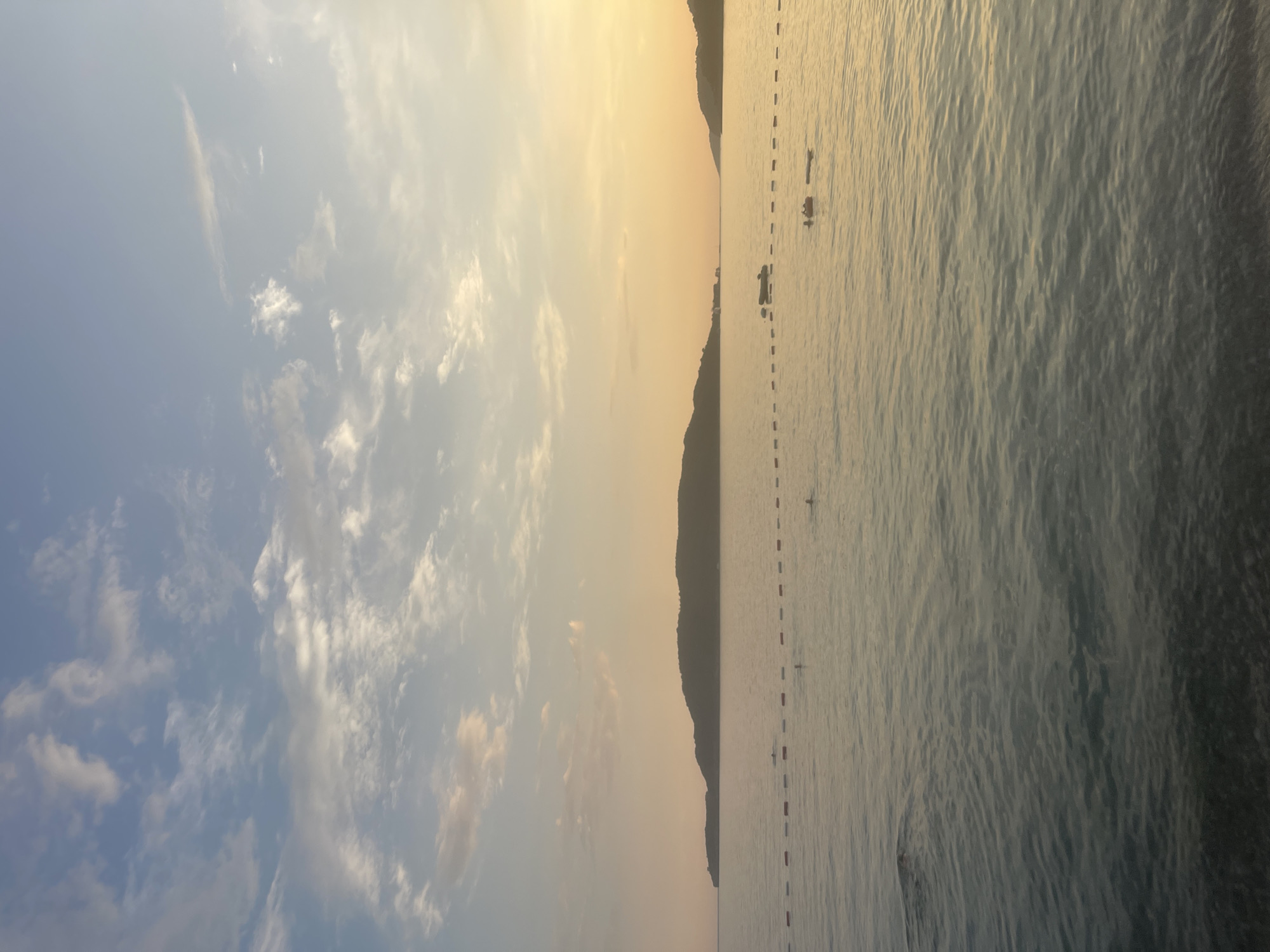 This is an image of the sea in Rafailovići, Montenegro. The sky is partly cloudy, with a blend of blue and pastel colors, indicating either sunrise or sunset. In the foreground, the water is calm with gentle ripples, and the seabed is visible in some areas. A line of buoys separates the swimming area from the open water. Several people are visible swimming and relaxing in the water. In the mid-ground, a long, hilly island or peninsula stretches across the horizon. There are also a few small boats or rafts in the water. The overall scene evokes a peaceful and serene atmosphere.
