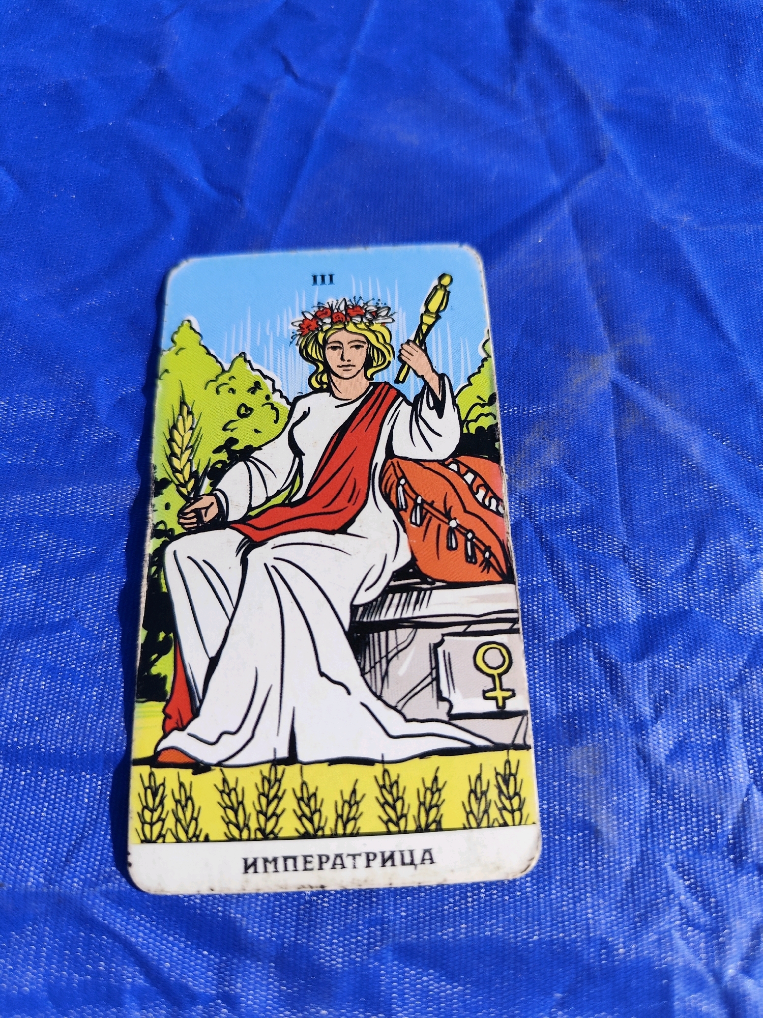 A tarot card, identified by the Russian text "ИМПЕРАТРИЦА" (Empress) at the bottom, lies on a blue, textured surface. The card depicts a woman wearing a white robe and a red sash, adorned with a floral crown. She holds a scepter in her right hand and a sheaf of wheat in her left. Behind her, a stylized representation of trees and a bright sky is visible. To the right of the woman, a distinctive female symbol is present. The card itself is slightly angled. The blue surface on which it rests appears to be fabric with a repeating pattern. No specific location in Balqaş, Kazakhstan, can be determined from the image.