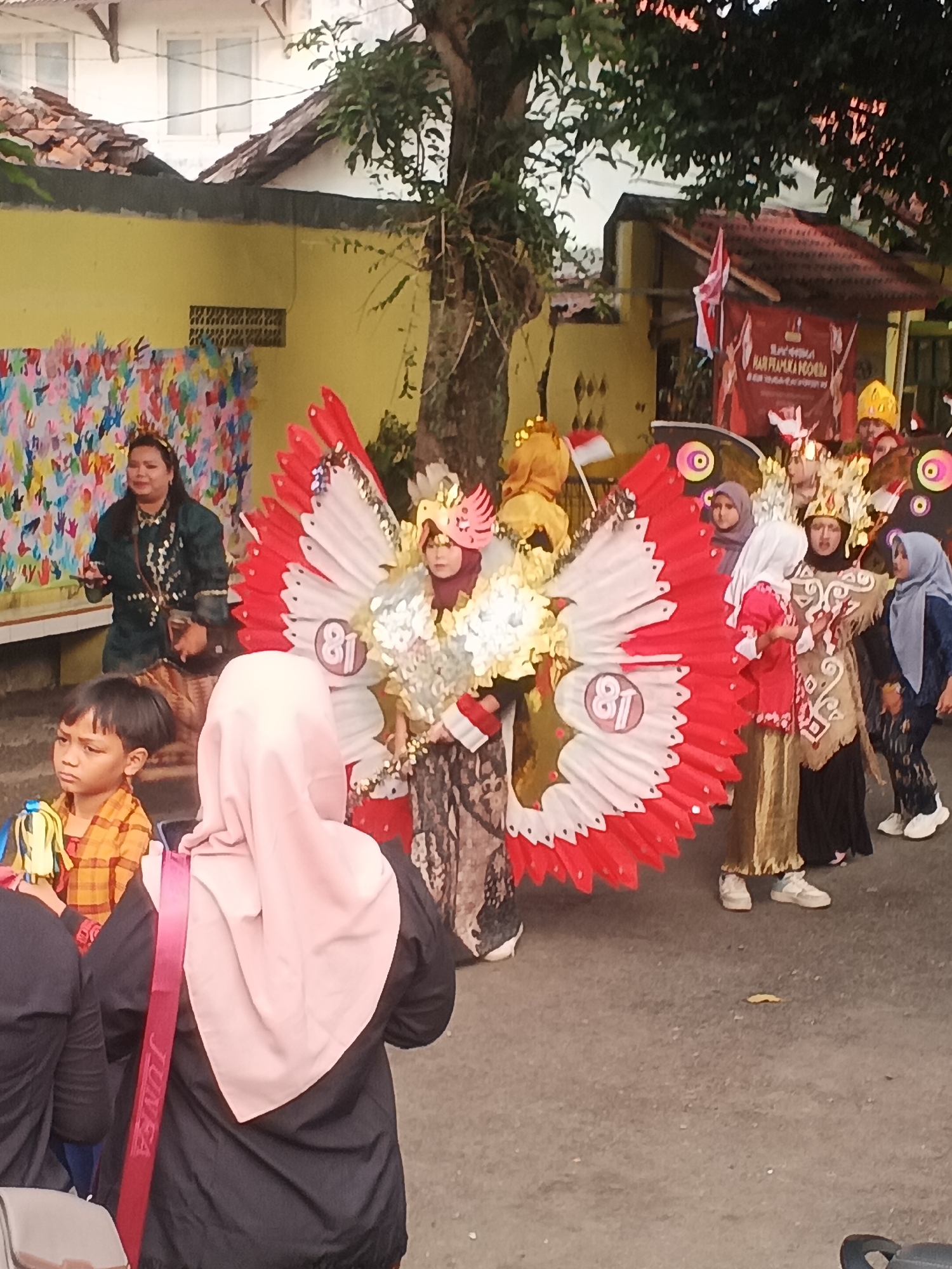 In Sokanegara, Indonesia, a group of people are participating in a parade. The foreground is dominated by a child wearing a yellow and orange plaid shirt, looking to the left. Behind the child, a woman in a pink hijab and a black jacket, with a red strap bearing the word "JUNEA", is visible. To the right of the child is a person in a large, elaborate costume resembling a bird with red and white wings. This costume has a prominent circular emblem with the number "81" on it. Other individuals are also in costume, with one person in a golden outfit adorned with a white pattern. Further back, a banner with Indonesian text and flags is displayed on a building. The environment appears to be a street lined with buildings and trees, suggesting a local celebration or event.