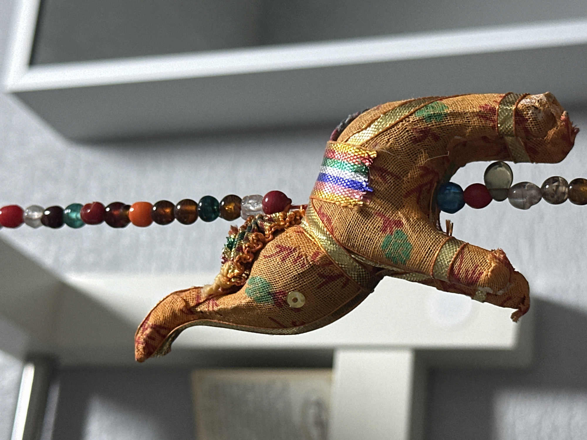 The image shows a close-up, detailed view of a fabric elephant ornament hanging from a beaded string. The elephant is predominantly orange with a woven texture and features a patterned fabric with red and green motifs. It is adorned with gold trim and a colorful striped ribbon around its back. A strand of multi-colored beads, including red, orange, brown, green, and clear, hangs above and partially to the side of the elephant. The background is out of focus, showing a light gray wall and a hint of a white frame, suggesting an indoor setting. There is a faint shadow cast on the wall. The image does not contain any specific indicators or context that would allow for its localization to Nevinnomyssk, Russia.
