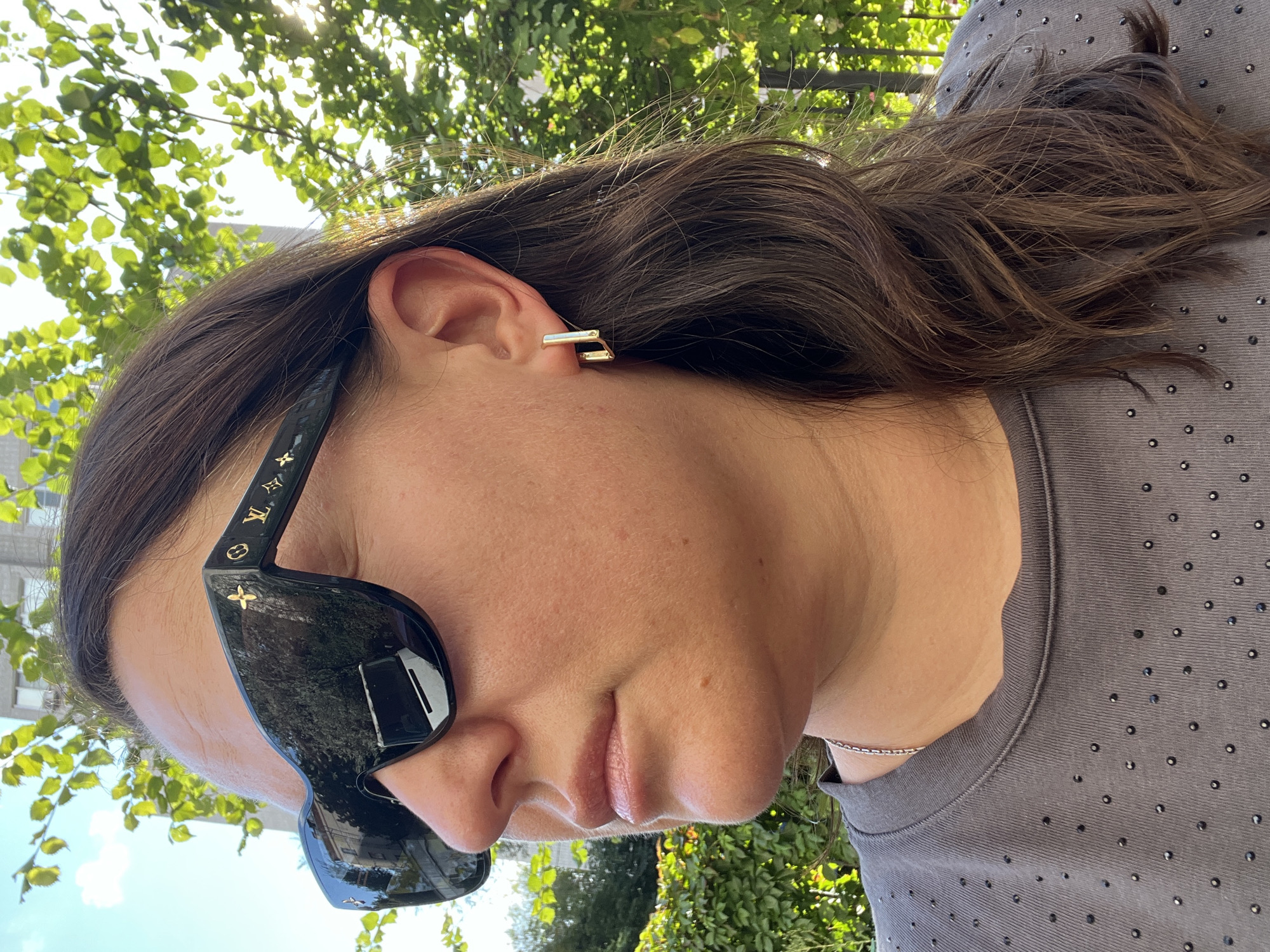 A woman is seen from the shoulders up, outdoors, with green foliage and a building in the background. She is wearing large, black sunglasses with a gold logo pattern on the frames. A gold, geometric earring is visible on her right ear. She is also wearing a dark gray t-shirt adorned with small, dark embellishments. Her brown hair is loose and falls over her shoulder. The lighting suggests it is daytime, with sunlight filtering through the leaves. The building in the background appears to be made of brick. The scene is likely a park or a residential area in Moscow, Russia.