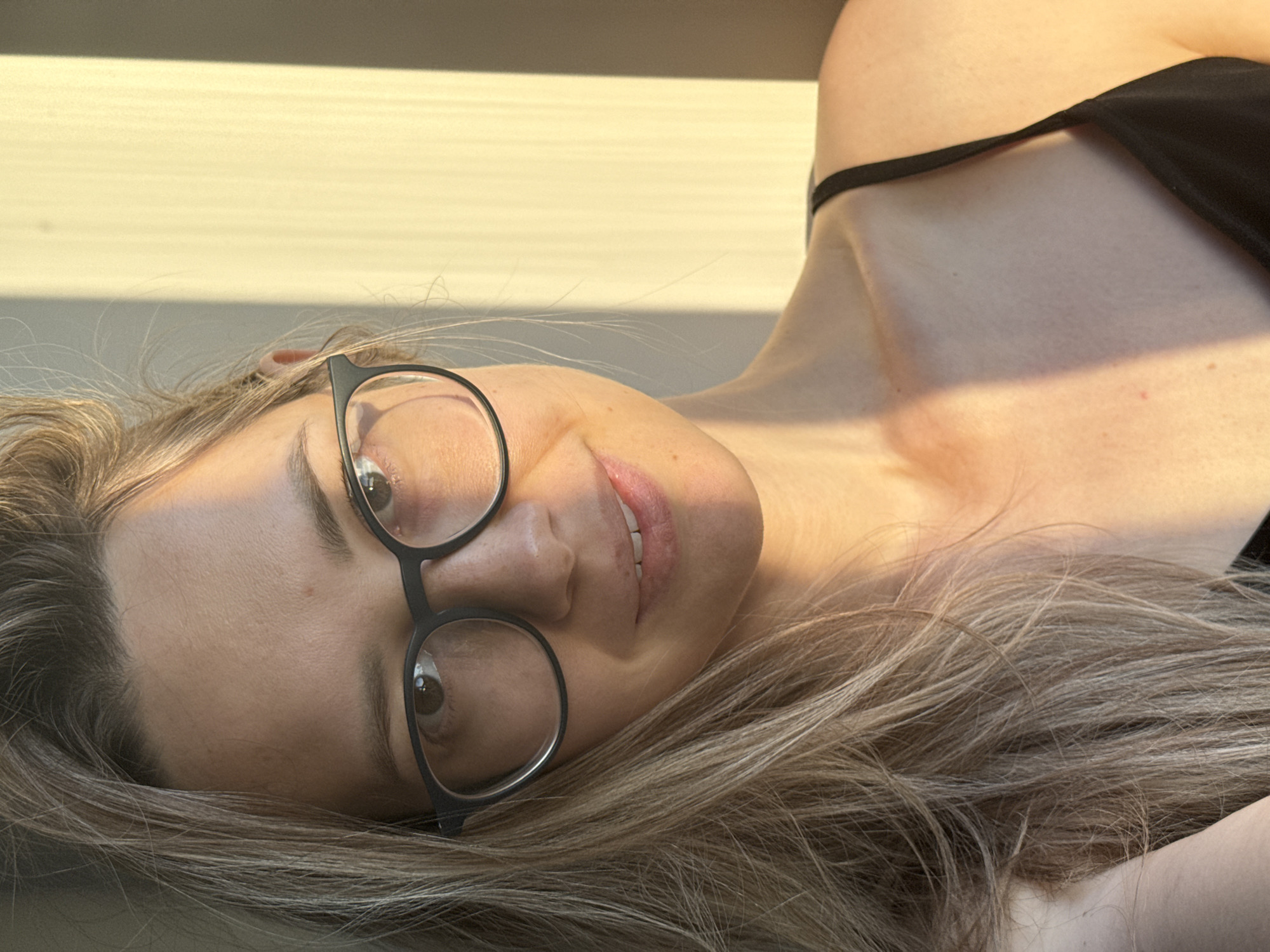 A woman with light brown hair is lying down, looking at the camera with a slight smile. She is wearing glasses with black frames and a black top with a thin strap. The background is a light yellow, possibly a wall. The location is not discernible from the image.