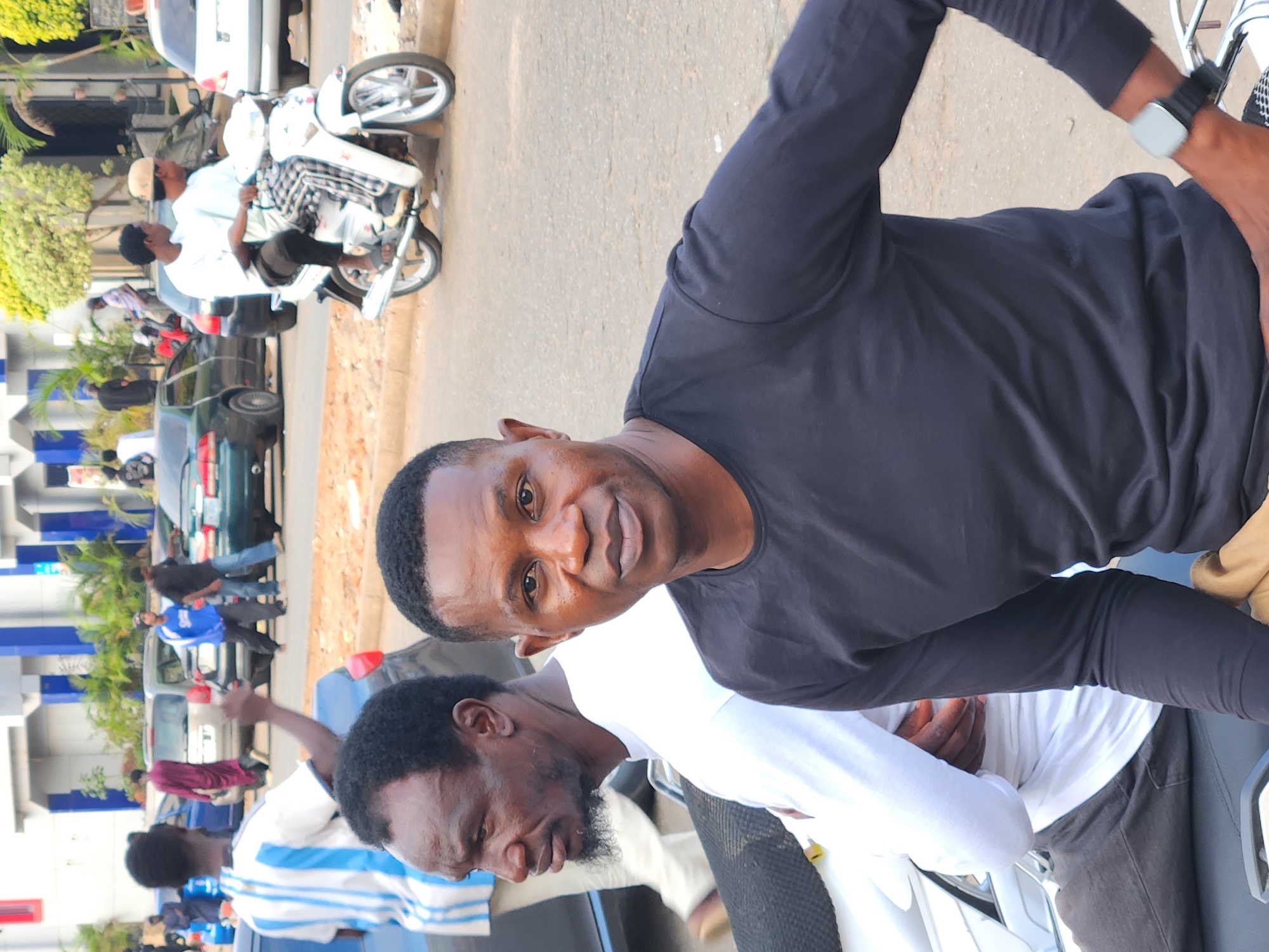 This daytime photograph, taken in Jos, Nigeria, captures a bustling street scene with two men prominently featured in the foreground. The man on the right, with dark skin and short dark hair, smiles directly at the viewer. He wears a black long-sleeved shirt and a light-colored smartwatch with a dark strap on his left wrist. He appears to be seated on a motorcycle or scooter, with a brown or tan garment visible near his waist.
Next to him, another man with dark skin, a dark beard, and short dark hair looks down and to his left. He is dressed in a white long-sleeved shirt, possibly featuring blue stripes on the shoulder. Both men are positioned close together, suggesting they are on the same vehicle or standing side-by-side.
The background depicts a vibrant streetscape typical of a Nigerian city. Numerous cars and other motorcycles are visible on the paved, light brown road. A white motorcycle with two riders, one wearing a light-colored cap, is distinct in the mid-ground. Buildings with blue and white accents, along with green trees and bushes, line the street, indicating an urban setting. Several other people are dispersed throughout the background, contributing to the lively atmosphere. The natural lighting suggests it is daytime. No discernible text is visible in the image.