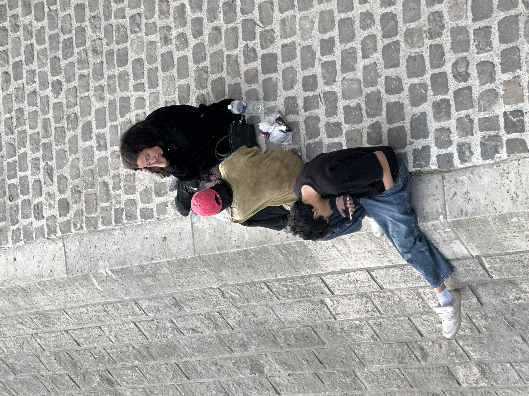 Three people are sitting on the ground next to a stone embankment. A woman wearing black is lying down with her eyes closed. Next to her sits another person wearing a mustard-colored shirt with a red hat next to them. The third person, wearing jeans and a black top, is holding a phone and looking down. A water bottle and a shopping bag are next to the woman. The location appears to be Paris, France, based on the architecture and stone embankment.