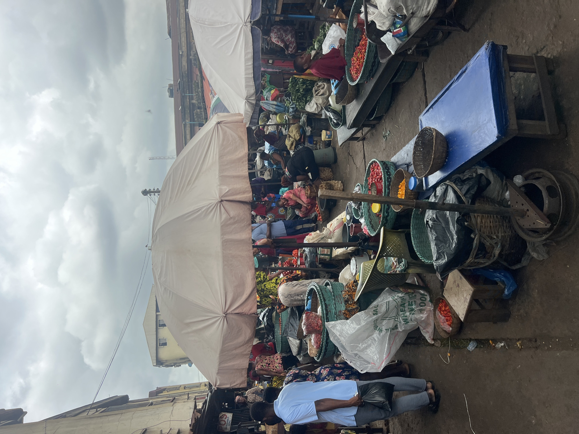 This is a photograph of a crowded outdoor market in Nkpor, Nigeria, on a cloudy day. The market appears to be a wholesale vegetable market, with numerous vendors selling produce, primarily tomatoes and peppers, in baskets and sacks. Large white umbrellas provide shade over some of the stalls. Several people are visible, some of whom are vendors attending to their goods, while others appear to be customers browsing or making purchases. The ground is wet and appears to be made of dirt or pavement. The overall impression is one of a busy, active marketplace.