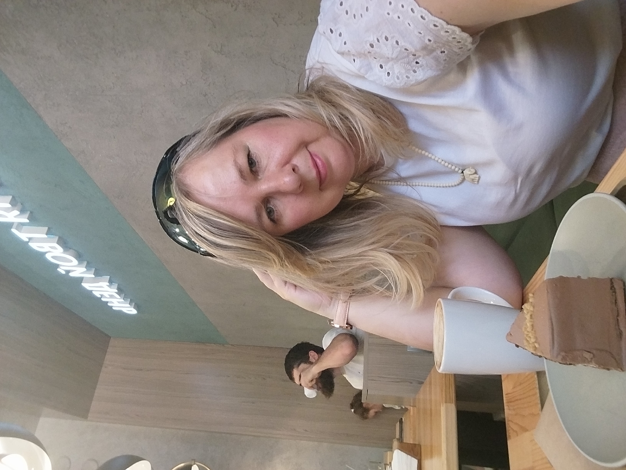 A woman with blonde hair and a white top is at a table with a cup of coffee and a slice of chocolate cake. A man with a beard is visible in the background. The wall in the background has "LEON DEHP" written on it. The picture was taken indoors, possibly in a cafe in Orenburg, Russia.