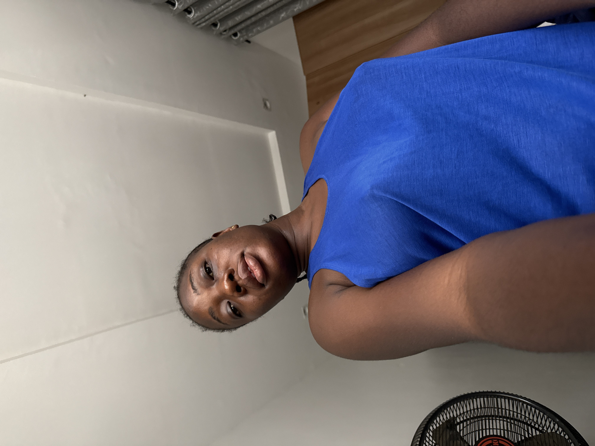 A woman is pictured in a low-angle selfie, likely taken indoors in Iddo Tudunwada, Nigeria. She is wearing a blue top. A white wall is visible in the background. Curtains are draped at the top left of the image, and a black fan is visible in the lower right corner. The woman is looking at the camera with a neutral expression.