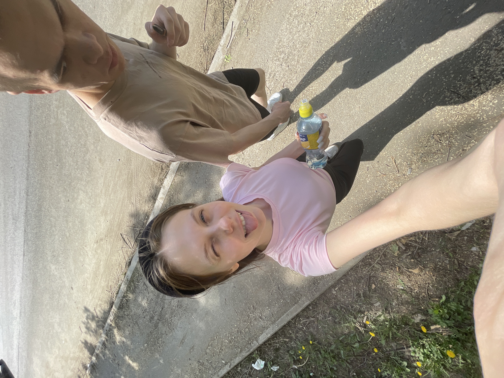 A high-angle photograph depicts two people standing on a paved path, likely in an urban environment. One person, appearing to be a male, is wearing a beige t-shirt and black shorts. The other person, appearing to be a female, is wearing a pink t-shirt and black shorts, and is holding a water bottle. The image is taken as a selfie, with the person in the pink shirt appearing to be the one holding the camera. The background consists of a concrete path and grassy area with yellow flowers. The location is presumed to be in the city of Ufa, Russia.