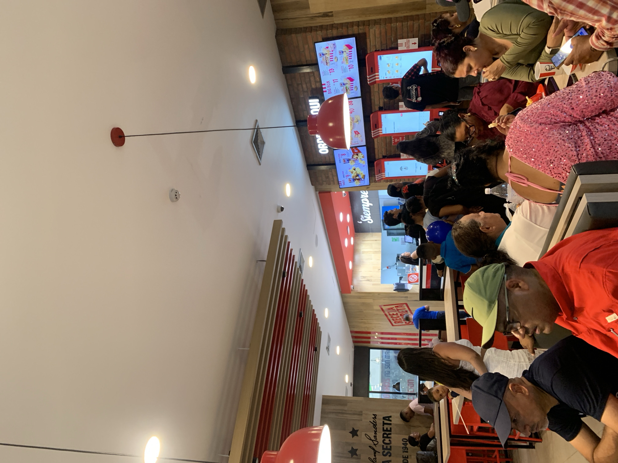 People are dining in a fast-food restaurant, possibly KFC, as suggested by the decor and menu boards. The scene is busy, with many customers seated at tables and seemingly enjoying their meals. Some are looking at their phones, while others are engaged in eating or conversing. The restaurant is brightly lit with multiple screens displaying menu items and prices.
Based on the text "RECETA SECRETA" and the location "land Sanders" visible in the image, it might be located in Porlamar, Venezuela. The environment includes red and white decorative elements and a large red light fixture.