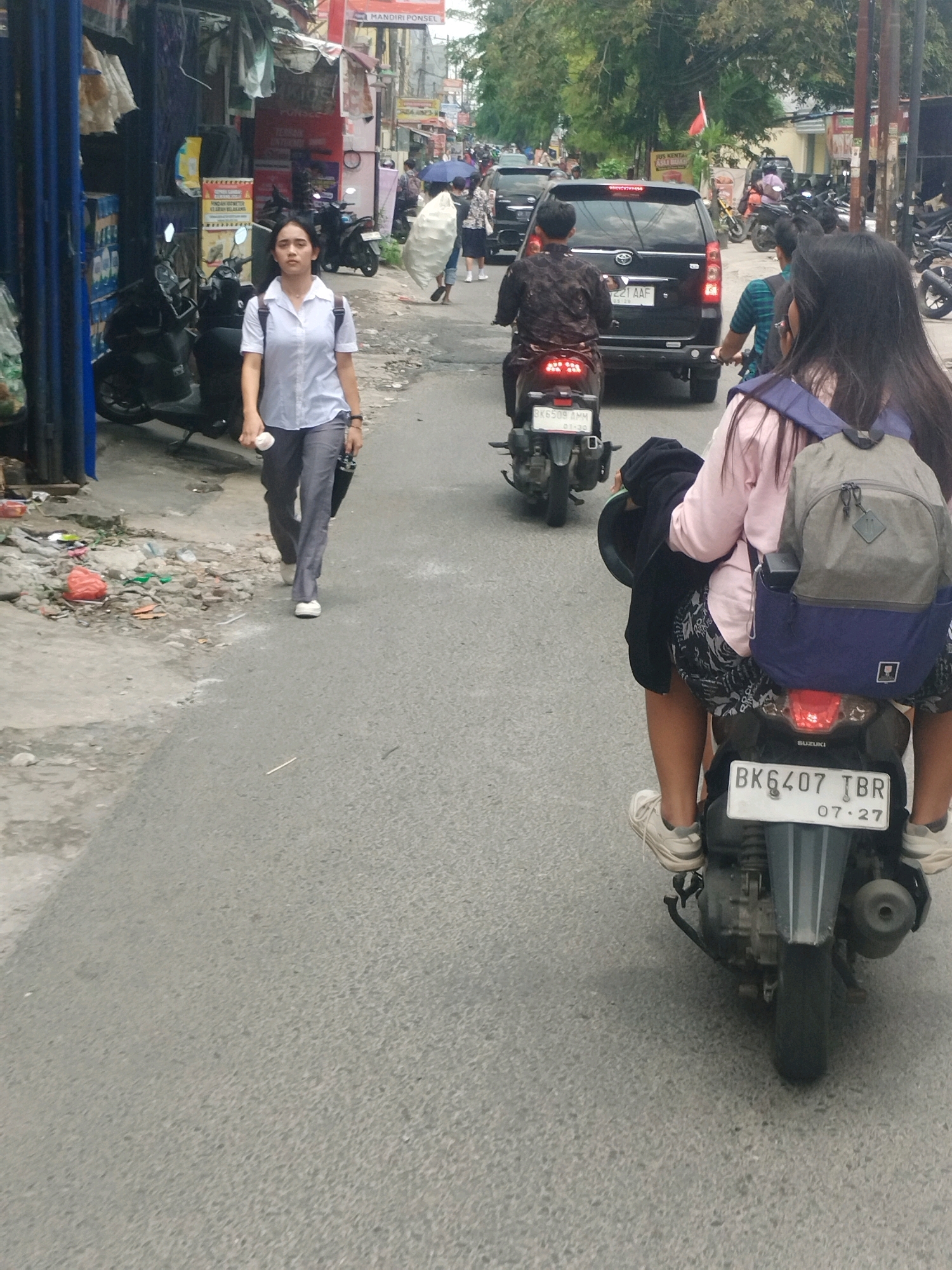 In the City of Medan, Indonesia, a street scene unfolds with multiple individuals and vehicles. On the left side of the road, a young woman walks towards the viewer, wearing a light blue collared shirt and grey pants. She carries a white object in her right hand and a black bag slung over her left shoulder. Several parked motorcycles are visible to her left, with shops and stalls lining the street behind her.
Further down the road, a black minivan with license plate "BK 221 AAF" is seen in motion. To the right of the minivan, two people ride on a scooter, with the person in front wearing a dark patterned shirt and the person behind, a young woman, wearing a pink shirt and a backpack. The scooter's license plate is "BK 6407 TBR". Other motorcycles and people are scattered throughout the scene, suggesting a busy urban environment. The road surface is asphalt, with debris and vegetation present along the edges. The background features buildings and trees, indicating an outdoor setting within the city.