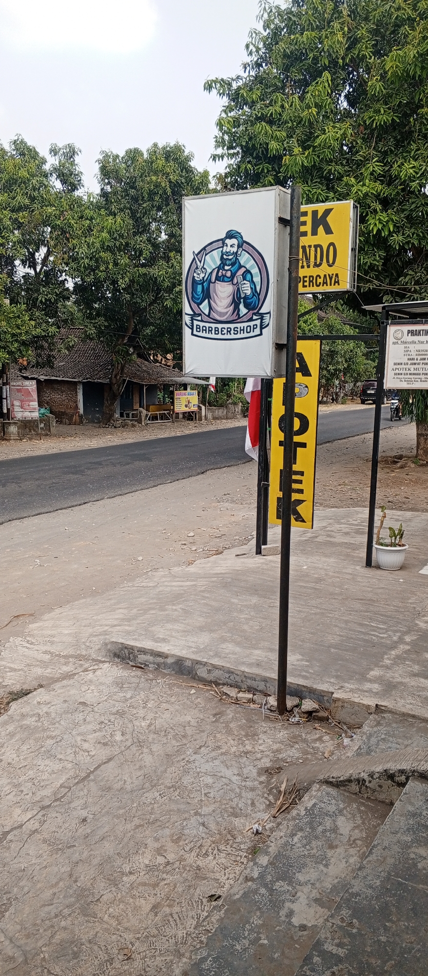 A barbershop sign with a cartoon barber is mounted on a pole on a street in Pingkuk, Indonesia. The sign is white with a blue and purple graphic of a barber holding scissors and a comb. Text below the graphic reads "BARBERSHOP". Next to the barbershop sign are two yellow signs, one with text that is partially visible. A building with a traditional Indonesian roof is visible in the background, along with trees and a road. A white pot with a green plant is on the sidewalk. The scene appears to be in a rural or semi-rural area.