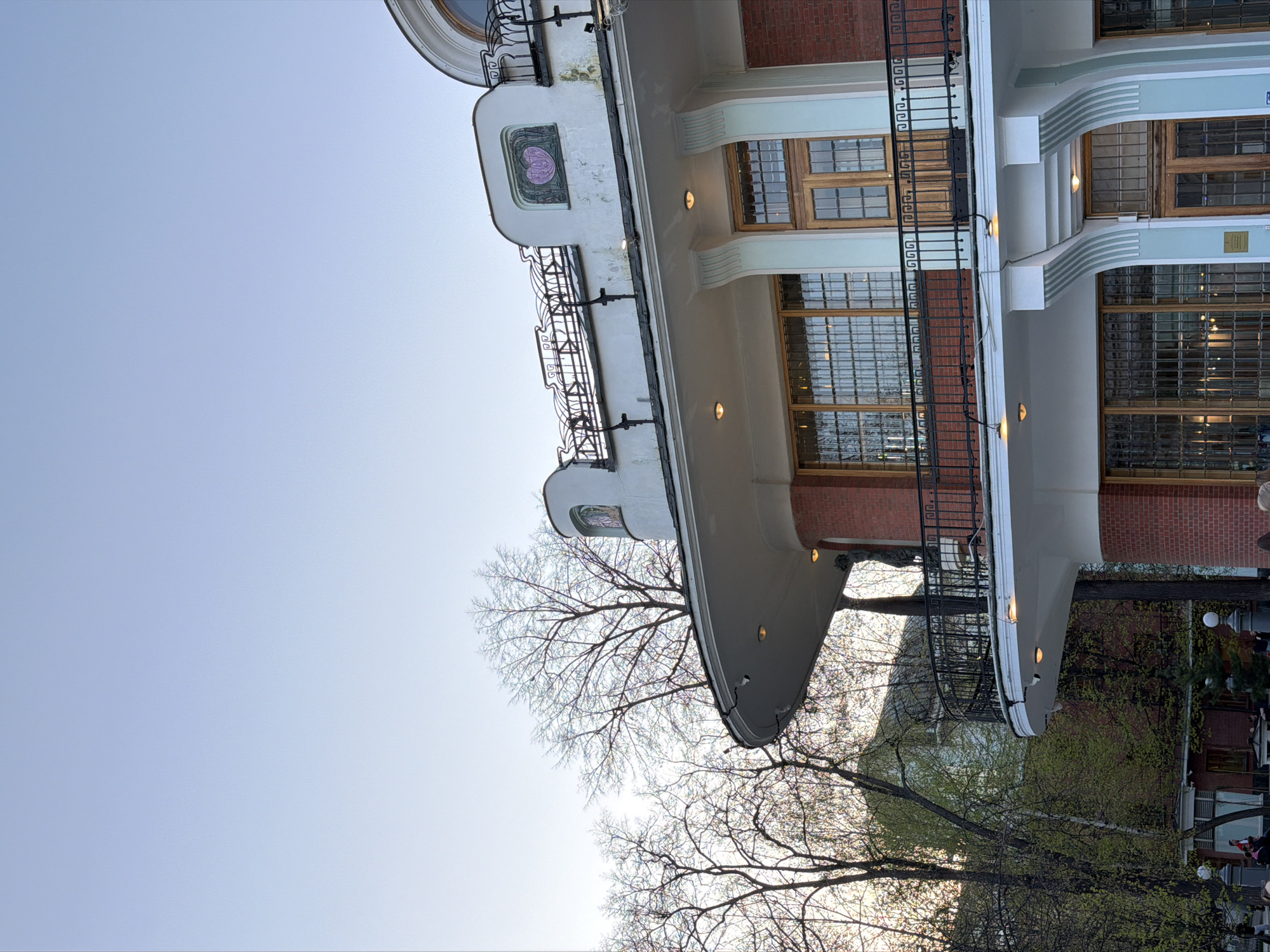 The image captures a unique architectural structure against a pale blue sky. The building features a curved, multi-tiered design, with sections adorned in white, light blue, and red brick. Numerous windows with wooden frames are visible, along with intricate black iron railings. At the base of the building, bare tree branches and foliage are partially illuminated by sunlight. A few indistinct figures are present at ground level, suggesting people interacting with the building or the surrounding environment. This is likely a building located in Moscow, Russia.