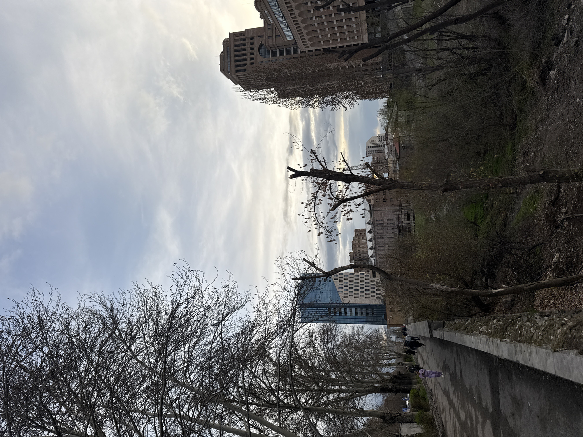 The image depicts a view of Yerevan, Armenia, likely from a park or elevated walkway. Tall buildings are visible in the background and among the trees. The sky is overcast with wispy clouds. Several bare trees are present in the foreground. Along a paved path, a few people can be seen walking in the distance. The camera is oriented at an oblique angle, with the ground appearing on the right side of the frame.