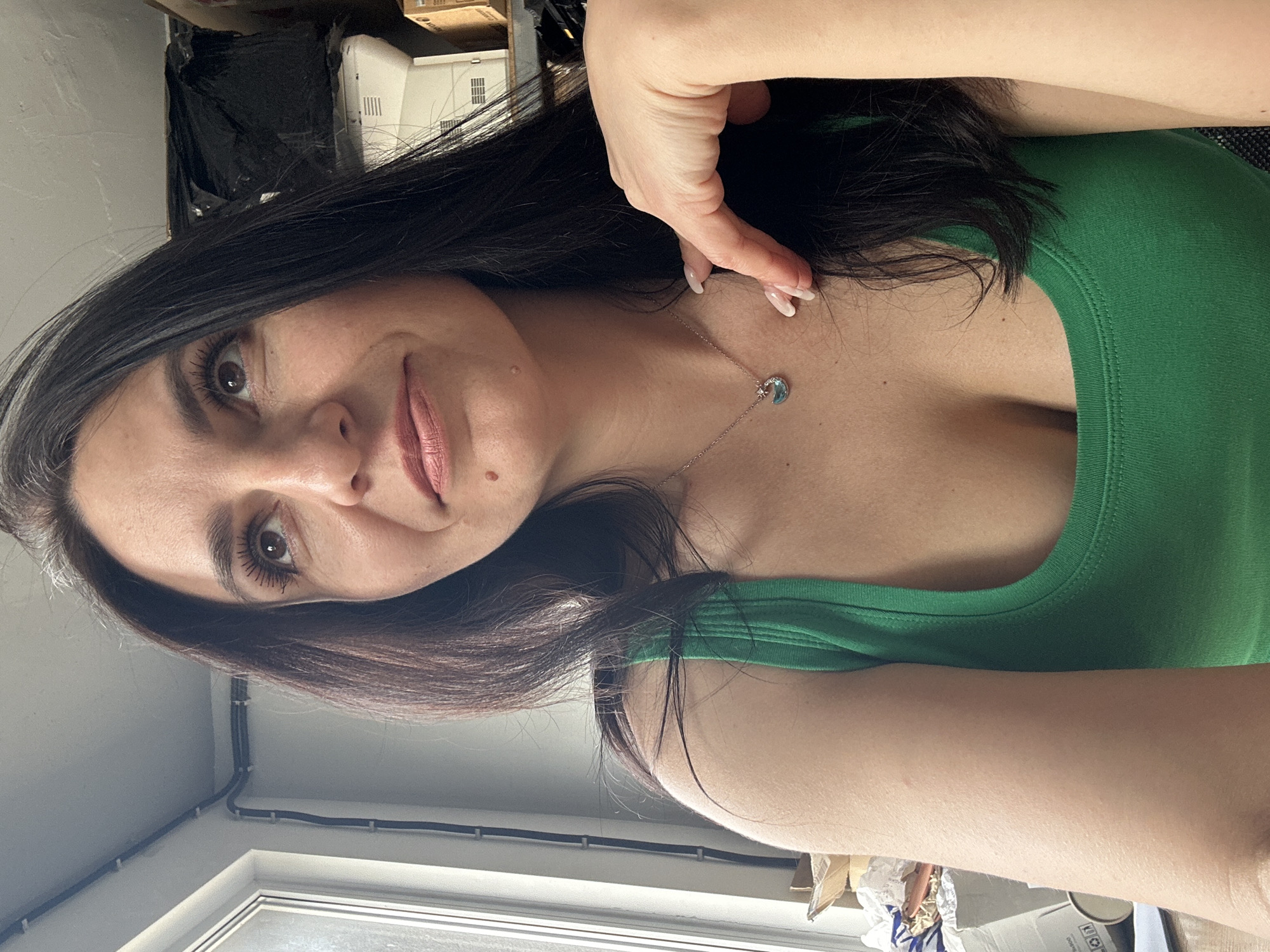 A woman with dark hair is seen from a close, elevated angle, wearing a green top and a necklace with a crescent moon pendant. Her face is turned slightly, with her left hand near her neck. The background includes a window with some electrical conduits, a wall, and some miscellaneous items on a table or shelf behind her, including some boxes, a black bag, and a white electronic device. The location appears to be indoors, but the specific city and state are not identifiable from the image.
