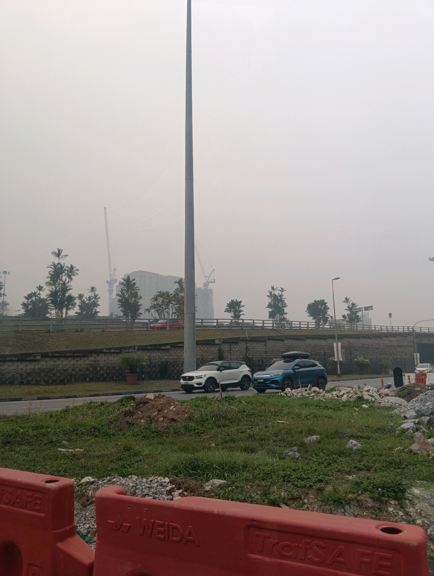 In a hazy urban environment in Kuching, Malaysia, a tall utility pole dominates the center of the frame. In the foreground, two large red traffic barriers, one with "SAFE" embossed on it and another with "WEIDA TrafSAFE" embossed, sit partially obscuring a patch of green grass and rocky debris. Beyond the barriers, a paved road is visible with two vehicles: a white SUV and a blue SUV with a rooftop cargo carrier. The white SUV is closer to the foreground, while the blue SUV is further down the road. A stone retaining wall runs horizontally across the middle ground, topped with a wooden fence. Behind the fence, palm trees and other vegetation are sparsely scattered. In the background, under a smoggy sky, construction cranes are visible, and a large building is under construction, suggesting ongoing development. The overall atmosphere is characterized by a thick haze, indicating poor air quality.
