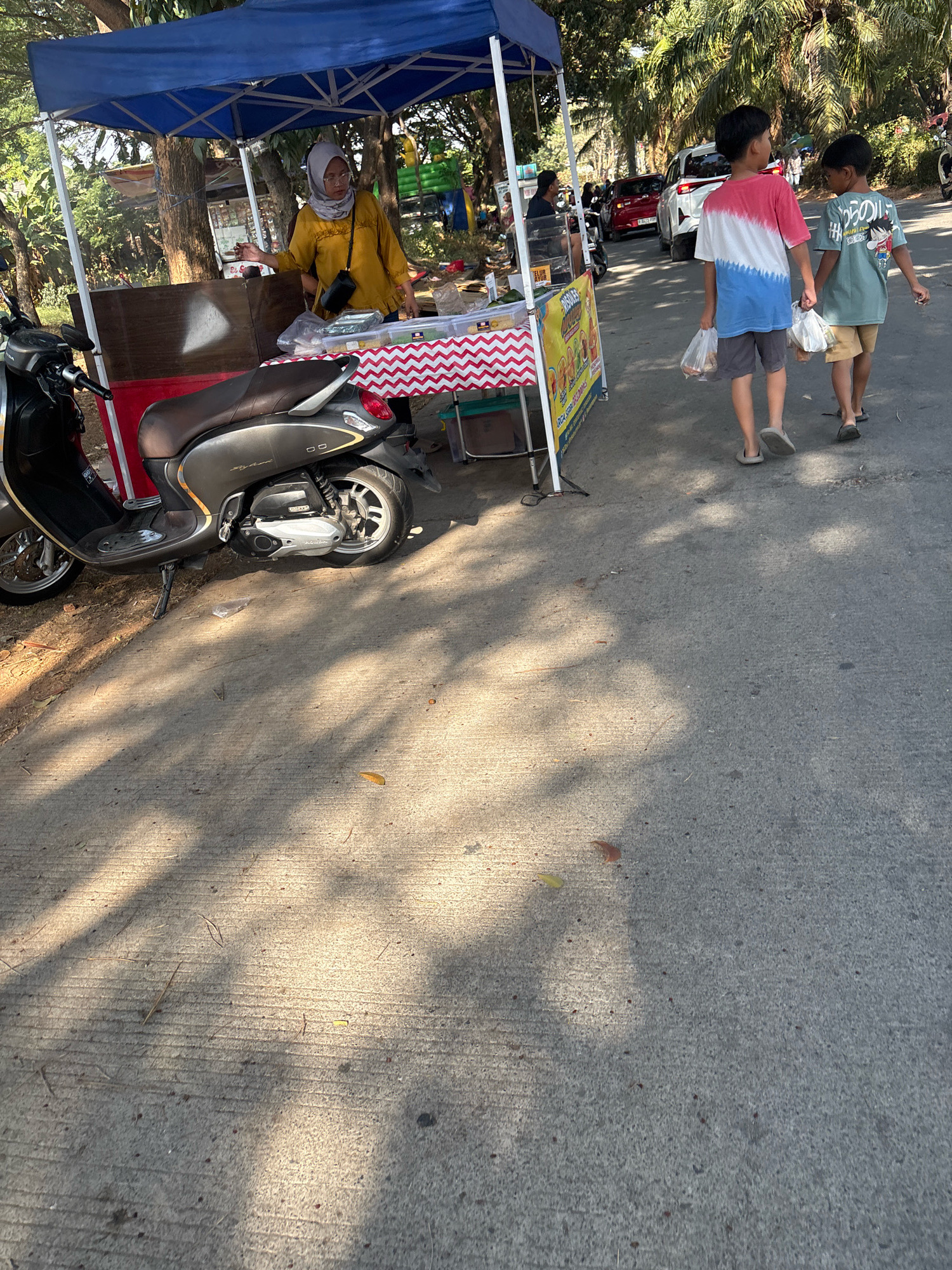 A street scene in Telagamurni, Indonesia, shows a woman in a yellow shirt and hijab standing behind a food stall. The stall is covered by a blue canopy and decorated with a red and white striped cloth. A scooter is parked to the left of the stall. Two young boys, holding plastic bags, walk away from the stall towards the right of the frame, passing by a white SUV and other cars on the road. Palm trees and other foliage line the street, casting dappled shadows on the concrete pavement.