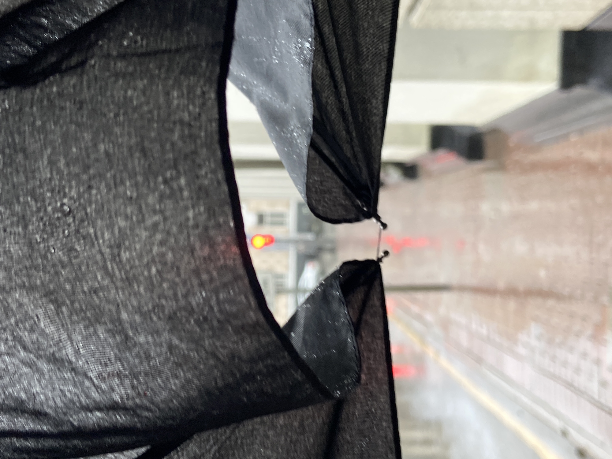A blurry view shows rain falling on a road in Moscow, Russia, as seen through a black fabric draped over a window. A red traffic light is visible in the distance. The road is wet, reflecting the red lights of vehicles. A brick structure lines the side of the road. The fabric is gathered in the foreground, partially obstructing the view.