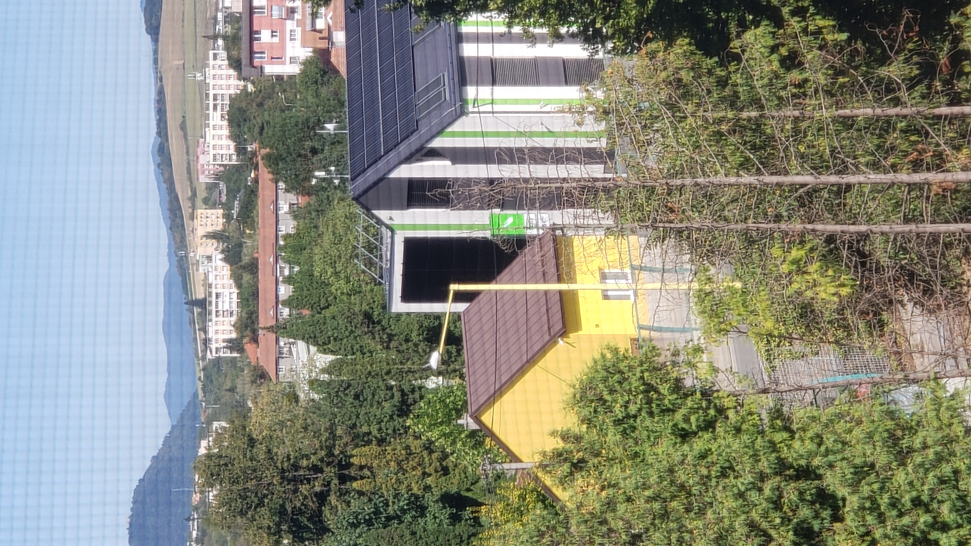 The image depicts a suburban or semi-rural landscape in Prešov, Slovakia, characterized by a mix of residential buildings, natural vegetation, and infrastructure. In the foreground, a bright yellow single-story house with a brown roof is partially visible, situated amidst dense green trees. Adjacent to this yellow house is a more modern, multi-story structure with a grey and white striped facade, featuring solar panels installed on its roof. This contemporary building also has large dark windows and a prominent green logo or sign.
Behind these two primary structures, a sprawling cityscape unfolds. Numerous apartment buildings, varying in color from white and yellow to pink, are spread across the midground. Some of these buildings appear to be mid-rise apartment blocks, suggesting a densely populated residential area. The background is dominated by rolling hills and mountains, some of which are covered in lush green vegetation, while others show signs of agricultural fields or lighter-colored terrain. The sky is a clear, pale blue, indicating fair weather. The overall impression is of a community nestled within a scenic natural setting, integrating modern residential development with the surrounding landscape. No people are visible in the image.