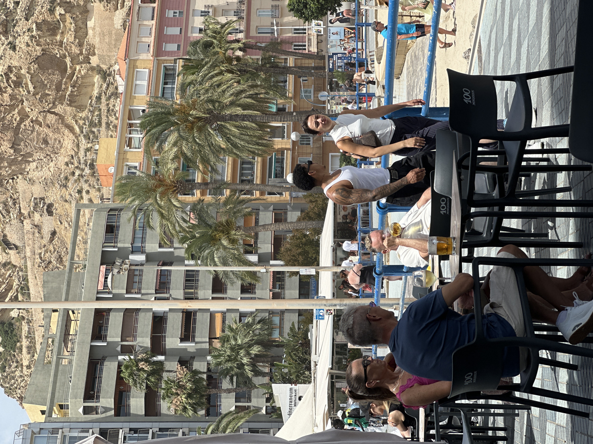 The image shows an outdoor seating area at a restaurant or cafe, likely in a Mediterranean coastal city given the palm trees and architecture. In the foreground, two people, a man and a woman, are seated at a table. The man, wearing a blue t-shirt and light-colored shorts, is holding a glass of beer. The woman, wearing a pink top, is seated next to him. Another man is visible in the background, drinking from a beer mug. Further in the background, several other people are walking around, and a blue railing separates the seating area from a sandy beach or promenade. Behind the people, there are buildings of various colors and heights, with a rocky hillside in the distance. The scene appears to be on a sunny day.