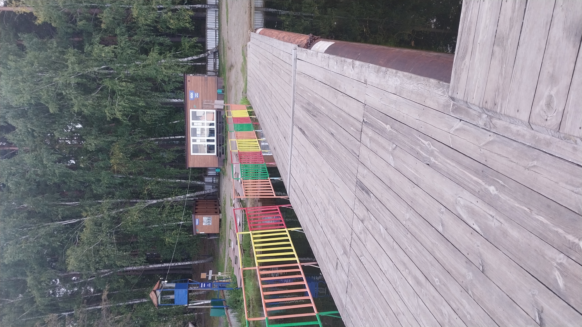 A wooden pier extends into dark water. A multi-colored fence, with posts colored red, yellow, green, and orange, runs along the edge of the pier. In the background, there is a wooden building with a white-framed window and a sign that says "КАФЕ". A small blue lifeguard tower stands to the left of the building. A dense forest of birch and pine trees borders the area. The scene appears to be a recreational area by a lake or river.