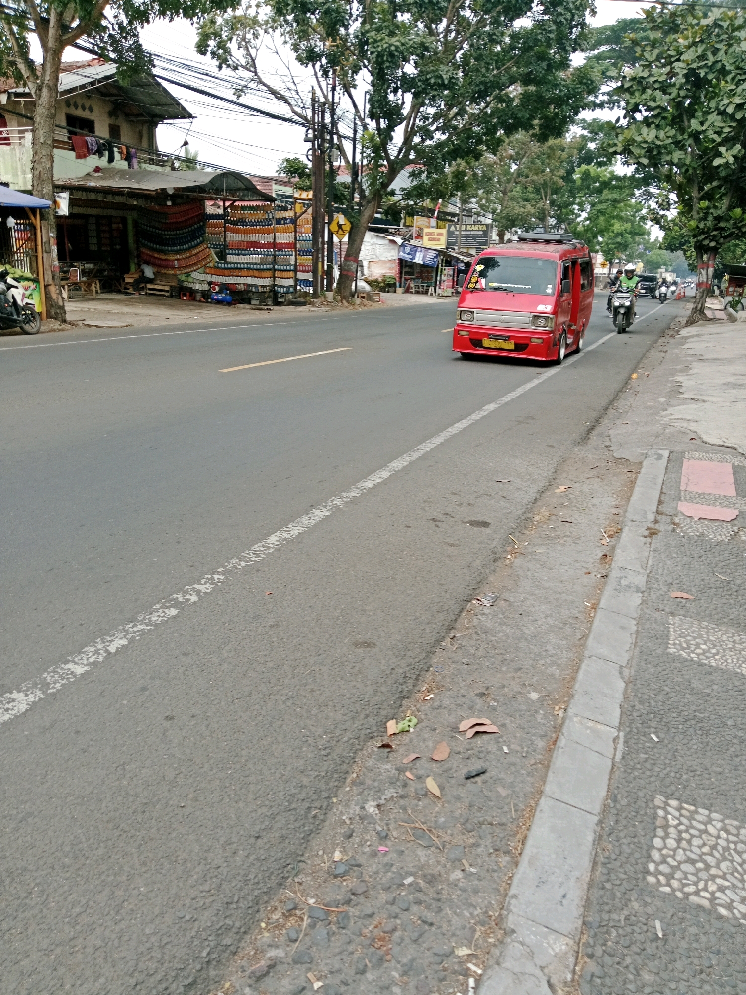 The image captures a street scene in the city of Muka, Indonesia. A red van is driving on the asphalt road, which has a white dashed line on the left side and a solid yellow line further into the distance. To the right of the road is a sidewalk with a concrete curb. The sidewalk is made of gravel and decorative stone patterns. On the left side of the road, there is a building with shops that display colorful items and a blue awning. Trees with green foliage line the street, casting shadows on the road and sidewalk. Several motorbikes are parked on the left side of the road near the shops. In the distance, more vehicles and buildings can be seen. The sky is overcast.