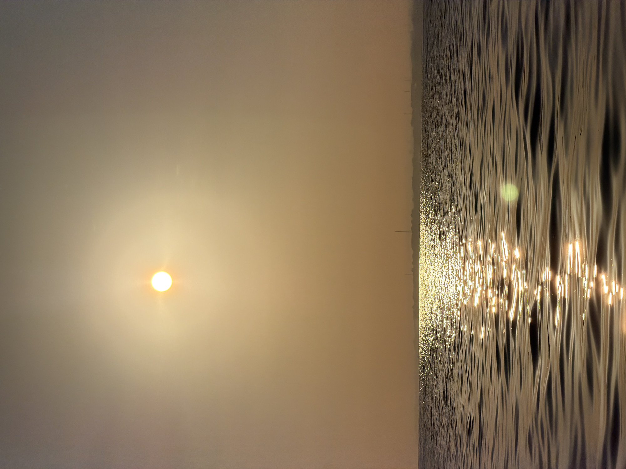 The image depicts a serene seascape at sunset or sunrise. The sun, a bright orb, is positioned high in the hazy, orange-tinted sky. Its light reflects intensely off the rippling surface of the water, creating a shimmering pathway towards the horizon. In the distance, a faint, silhouetted shoreline is visible, marked by industrial structures and possibly a few trees. The water itself is calm, with gentle waves creating patterns of light and shadow. The overall atmosphere is tranquil and atmospheric, suggesting a peaceful moment in nature.