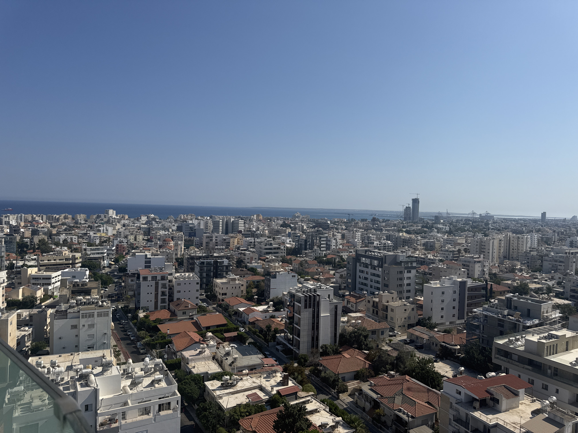 This is a high-angle, wide shot of a densely populated urban landscape, likely Limassol, Cyprus, looking towards the coast under a clear blue sky. The foreground shows the rooftop of a modern building with white solar water heaters and glass railings.
The midground is filled with a dense collection of apartment buildings and houses, characterized by flat roofs, many with visible solar water heaters, and some with red tile roofs. The buildings vary in height and architectural style, with many being multi-story structures in shades of white, grey, and beige. Streets are visible with parked cars, suggesting a residential and commercial area. Green trees are interspersed throughout the urban fabric.
In the background, the cityscape continues towards the horizon, with taller buildings and what appear to be port cranes and industrial structures in the distance, indicating a proximity to a harbor. The Mediterranean Sea is visible as a thin blue line separating the city from the sky. The overall impression is one of a bustling coastal city on a bright, sunny day. No people are clearly discernible in the image.