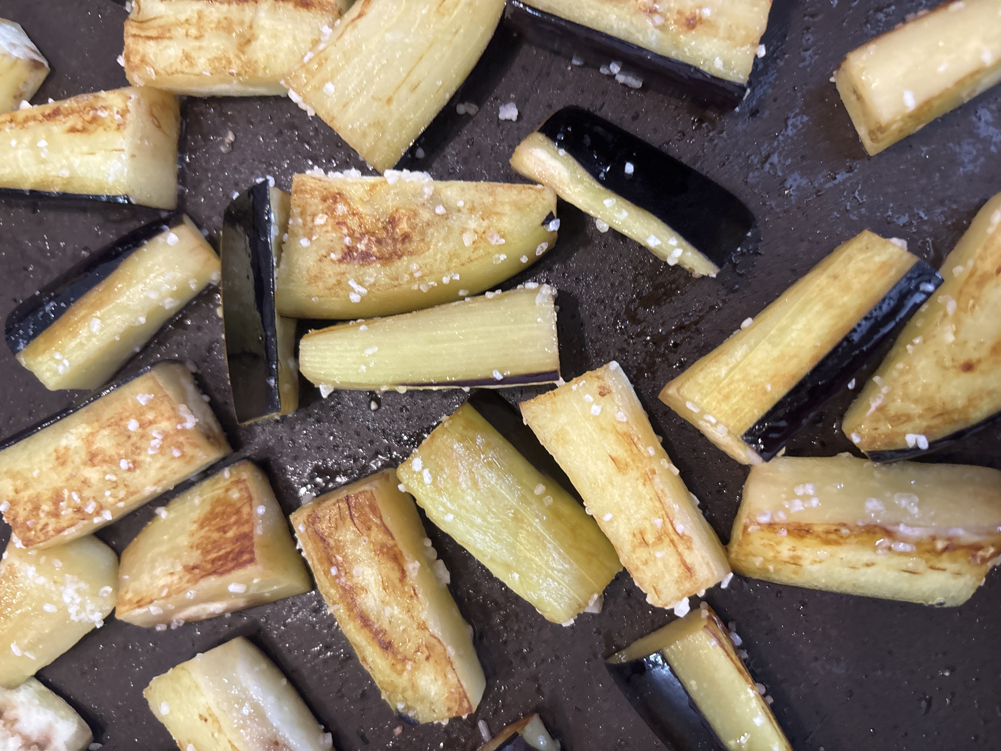 The image shows a close-up, overhead view of sliced and cooked eggplant pieces scattered on a dark surface, likely a baking sheet or pan. The eggplant pieces are cut into bâtonnet shapes and appear to be lightly browned and seasoned with coarse salt crystals. The overall color palette is dominated by the golden brown of the cooked eggplant and the dark background. There are no people or any indication of a specific location within Bila Tserkva, Ukraine. The scene is purely focused on the food item.