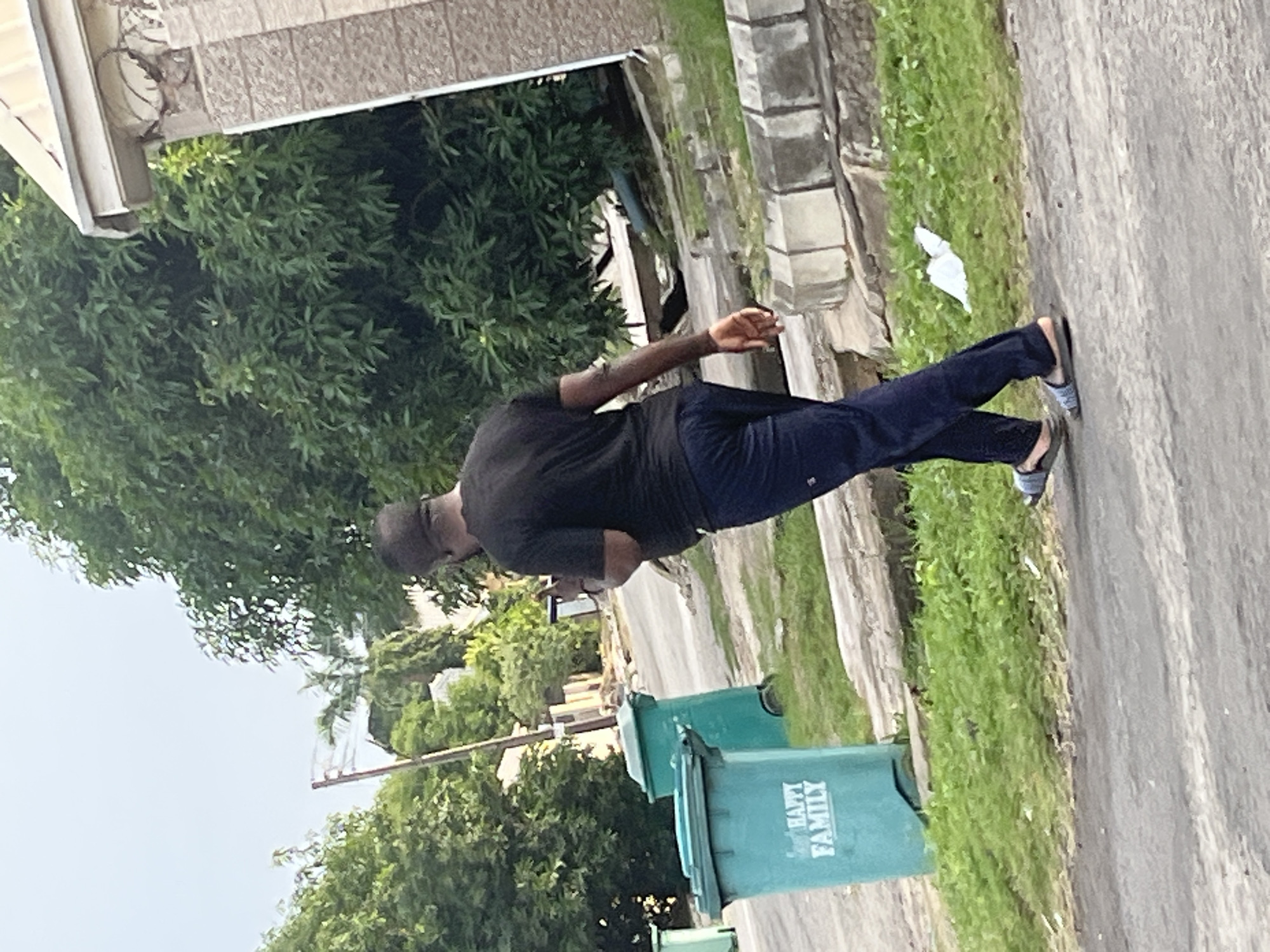 An adult male, appearing to be of African descent and wearing a black t-shirt and dark blue trousers, walks on a paved surface. He is holding a dark-colored mobile phone in his right hand. In the background, two teal-colored trash bins are visible. One bin has white text that reads "HAPPY FAMILY". The environment includes green grass, trees, and a building with a light-colored roof and stone facade. The scene suggests a street in Abuja, Nigeria, given the style of the buildings and vegetation.