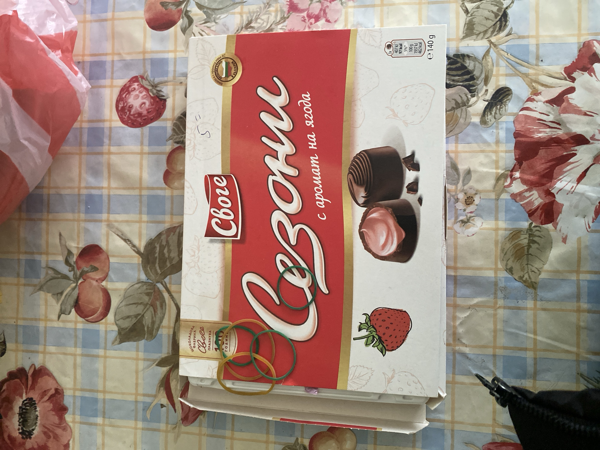 The image shows a box of "Sezoni" chocolates, produced by the "Svoge" chocolate factory in Bulgaria. The box features images of chocolate candies and strawberries, and states "with strawberry aroma". The background is a table cloth with a floral and plaid pattern. There are also several elastic bands on top of the box.