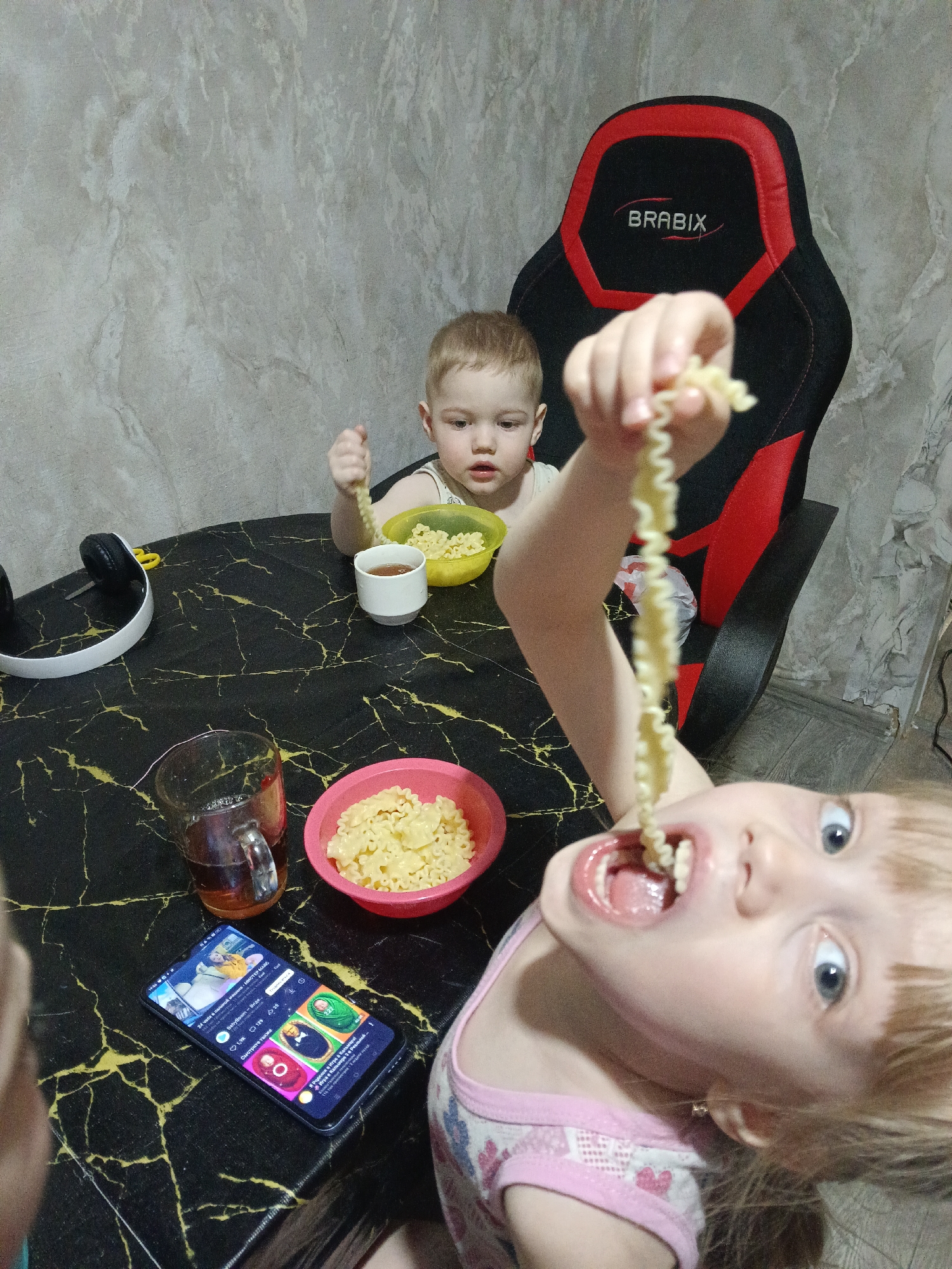 This image captures an indoor scene in Chaikovsky, Russia, depicting two young children at a table. The environment features a light gray, textured wall and a black table with prominent gold, marble-like veining. A black and red gaming chair with the "BRABIX" logo is visible in the background.
In the foreground, a girl, approximately 2-4 years old, with blonde hair and blue eyes, is wearing a pink and white sleeveless top. She is captured mid-action, holding a long, curly pasta strand with her right hand and inserting it into her widely open mouth, with her eyes directed towards the camera.
Behind her, to the left, a younger child, approximately 1-2 years old, with light brown hair and wearing a light-colored top, is seated in the gaming chair. This child holds a shorter, curly pasta piece in their right hand, positioned above a yellow plastic bowl filled with similar pasta. A small white cup containing a dark liquid is also in front of this child.
On the table, besides the children's items, there is a pink plastic bowl of pasta, a clear glass mug containing a dark liquid, a pair of white over-ear headphones with black earcups, and a small pair of yellow-handled scissors. A dark-colored smartphone lies face up, displaying a social media interface with Russian text and various visual posts, including images of babies and caricatures of older men. A crumpled white plastic bag is visible on the floor beneath the gaming chair.
