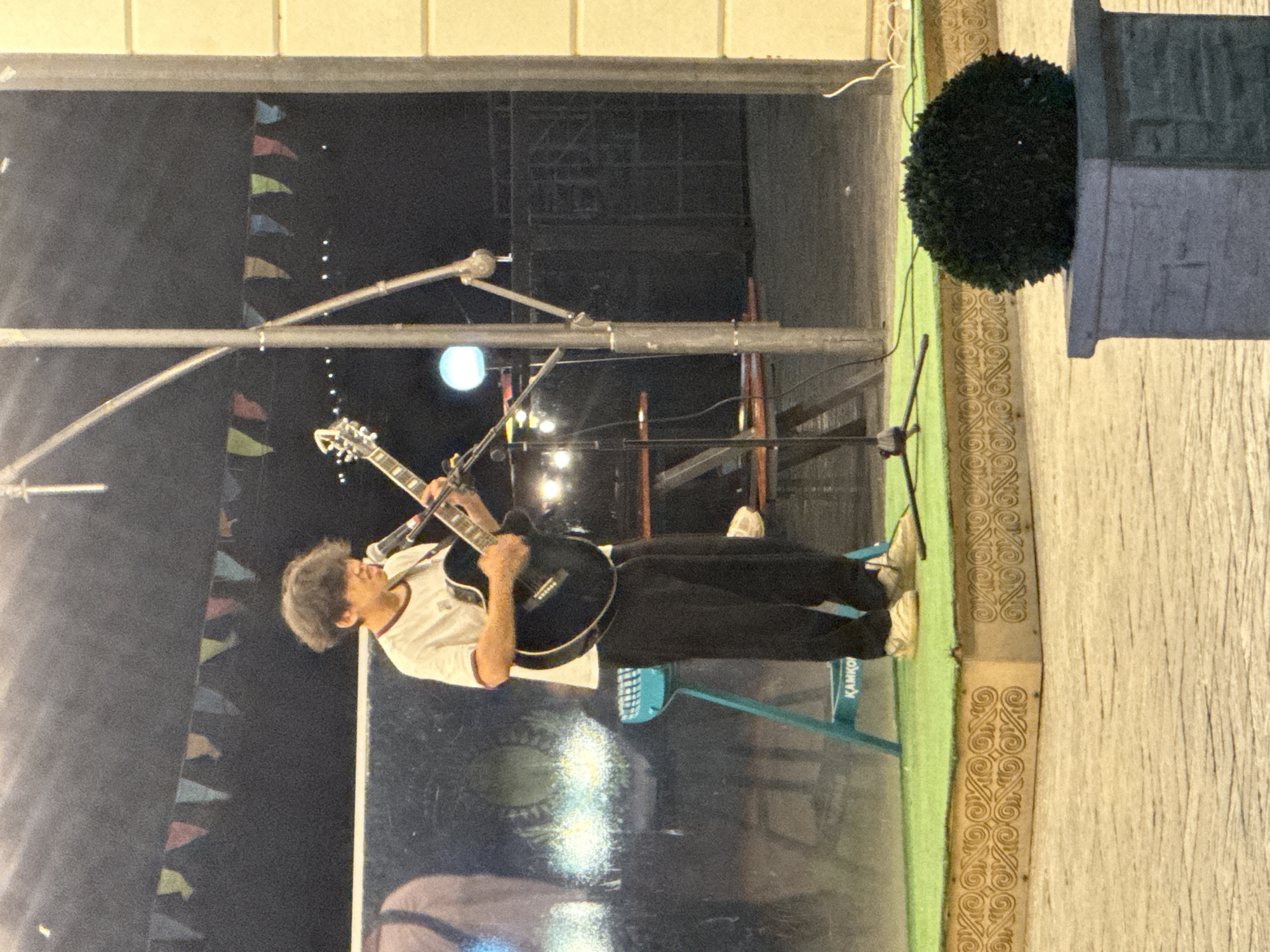 A person is playing an acoustic guitar on a stage at night, as indicated by the dark background and artificial lighting. The musician, with curly hair and wearing a white t-shirt and black pants, is focused on their instrument. The stage is adorned with colorful triangular flags, suggesting a festive or outdoor event. A microphone stand is positioned near the guitar. In the background, stage lighting and scaffolding are visible. To the right of the frame, a decorative, circular green shrub is present next to a textured, light-colored wall. The flooring of the stage appears to be dark and reflective, showing some light patterns. There is also a visible blue chair with the text "KAMKO" on it. The overall scene suggests a live musical performance during an evening event, possibly outdoors, in Pavlodar, Kazakhstan.