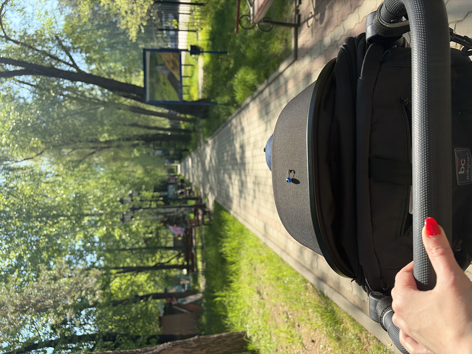 The image, taken in the city of Chaikovsky, Russia, shows a view down a paved pathway lined with trees and green spaces. A stroller is in the foreground, with a hand gripping the handlebar. The pathway extends into the distance, where benches and other park furniture can be seen. The perspective suggests someone is pushing the stroller along the path. A sign is visible along the path. The lighting suggests it's a sunny day.