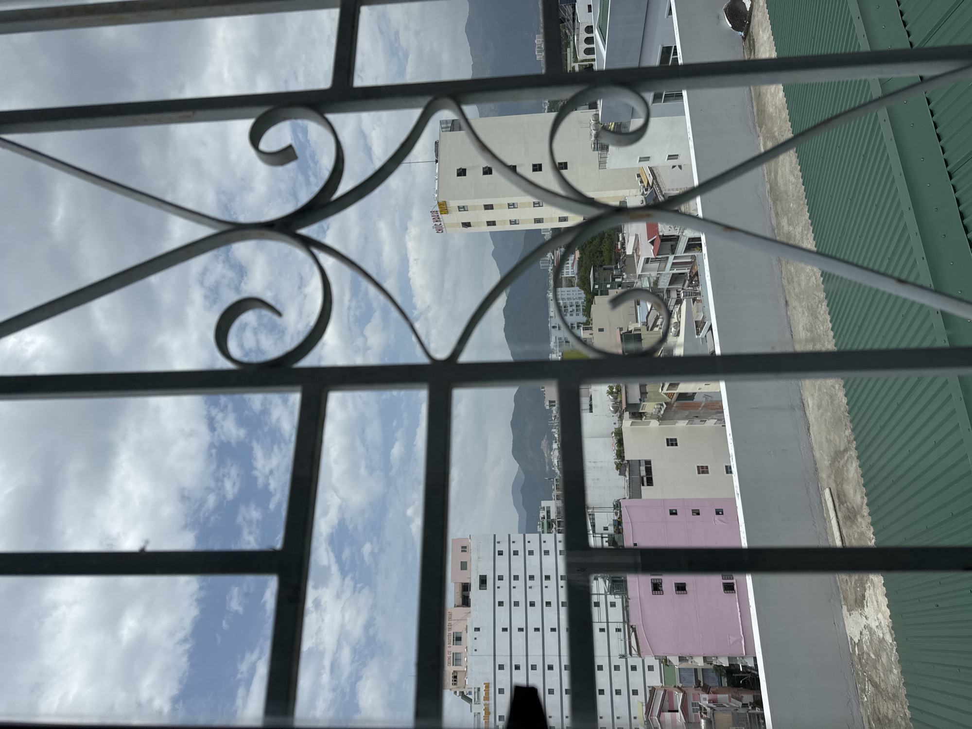 This is a photograph taken from behind a metal grate, looking out at a cityscape in Nha Trang, Vietnam. The view is dominated by buildings of various heights and colors, including a prominent white high-rise with "Sunlight Hotel" signage on its left side and another white tower with "CHIC HOUSE" and "HOTEL" signage. There are also pink and other colored buildings visible, along with rooftops and what appears to be a green corrugated metal roof in the foreground. In the background, mountains can be seen against a partly cloudy sky. The overall impression is of a dense urban environment with a backdrop of natural scenery.
