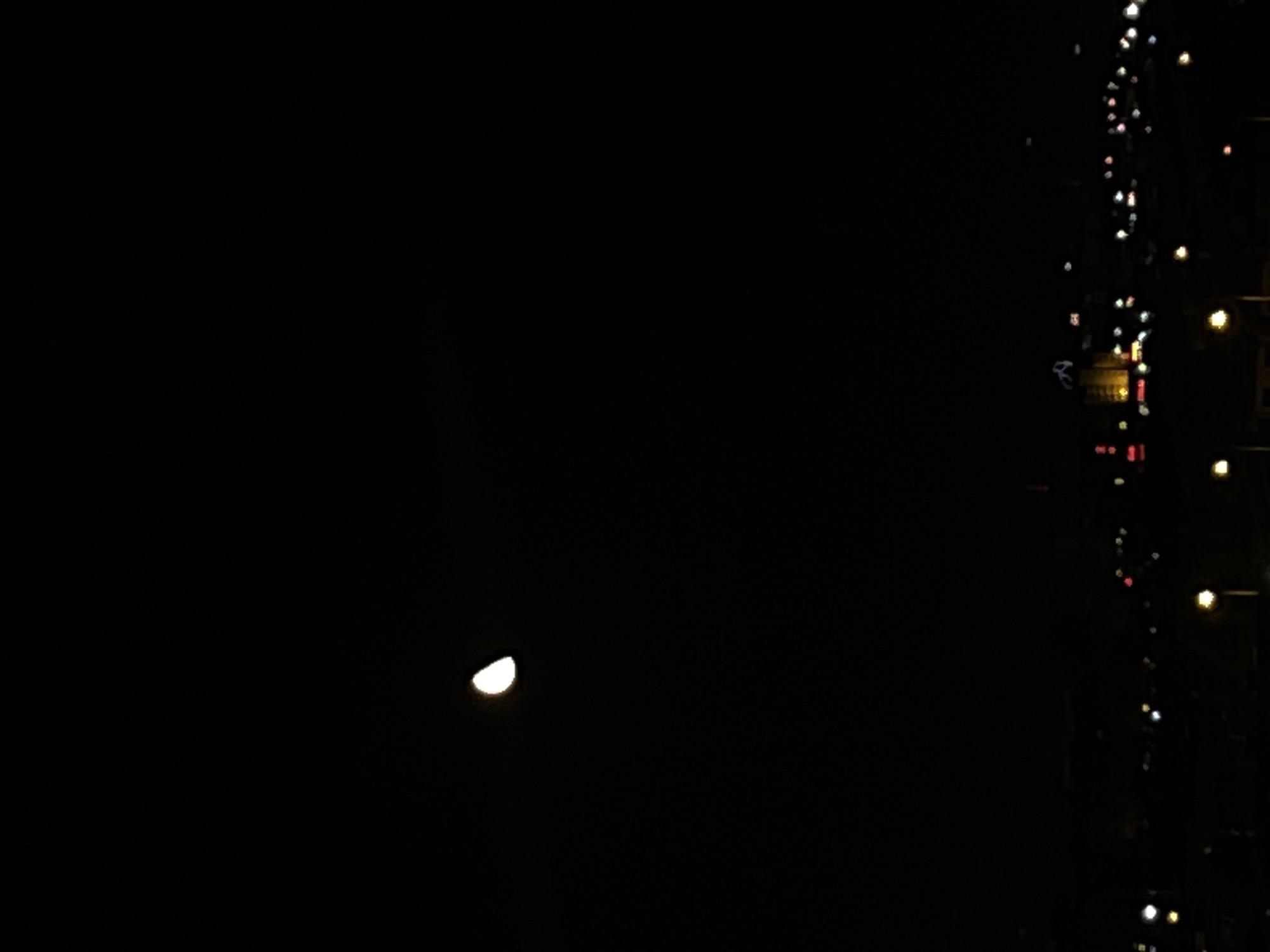 A night scene showing a crescent moon in the lower left quadrant against a dark sky. A vertical strip of blurred lights suggests urban infrastructure, possibly buildings and streetlights, in the right portion of the frame. The location is consistent with an urban setting at night, potentially Sofia, Bulgaria, given the information provided. No people are visible.