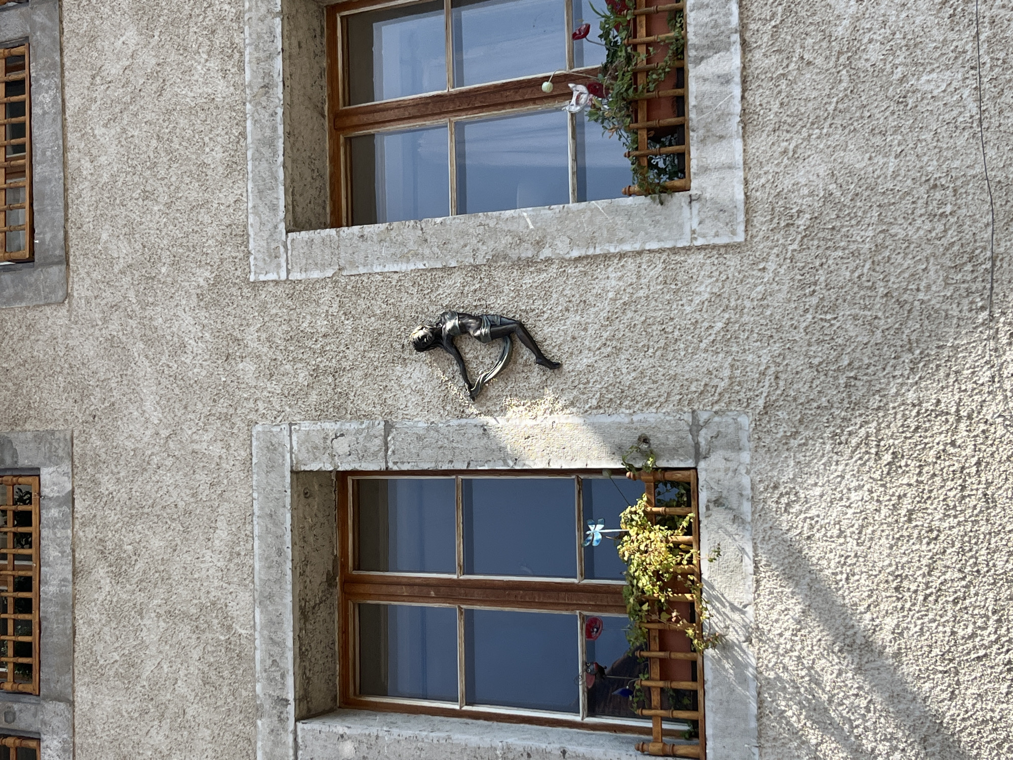 The image shows a textured, light-colored stucco building facade with two windows. The windows have wooden frames and glass panes. Above each window, there is a wooden grate. Below the left window, there is a wooden planter box with green foliage and two small decorative ornaments, one resembling a ladybug and the other a poppy. Below the right window, there is a similar wooden planter box with green foliage and some red flowers, along with a small white bird ornament. Mounted on the wall between the two windows is a dark-colored metal sculpture of a dancing female figure. The scene appears to be outdoors during daylight, with shadows cast on the wall from the left. The city of Epagny, Switzerland, is mentioned as the location, although no specific landmarks or identifying features of Epagny are visible in the image itself.