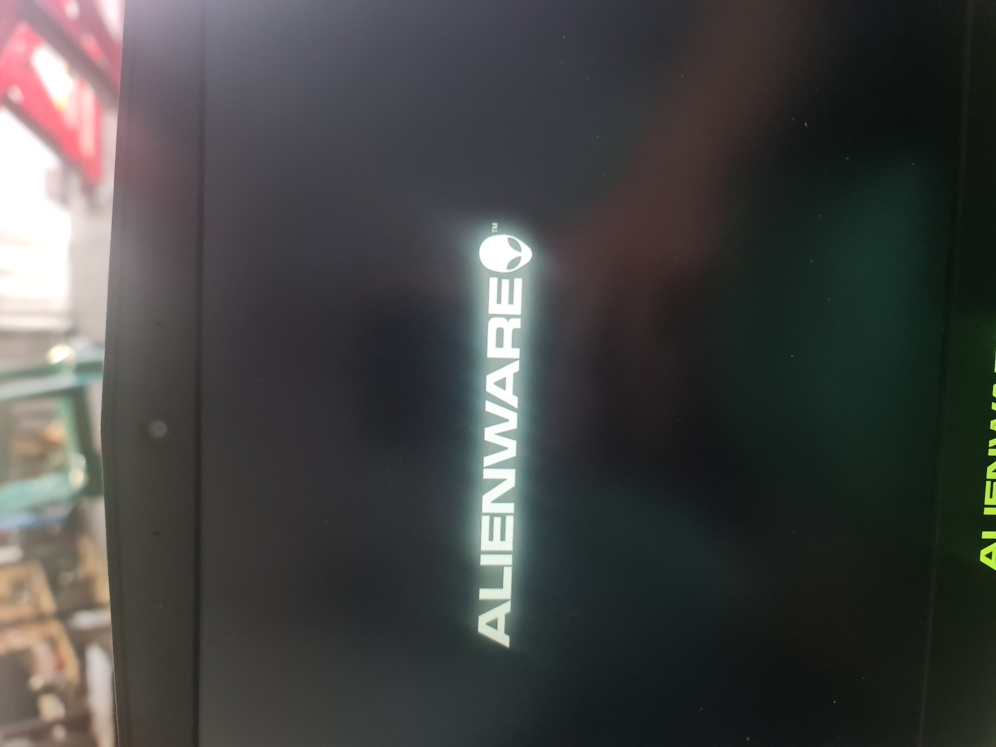 A computer screen is displayed vertically, featuring a dark background with the white "ALIENWARE" logo and its iconic alien head graphic prominently in the center. A small trademark symbol ("™") is visible near the alien head. The device appears to be in the process of booting up, as indicated by the brand splash screen.
On the left side of the screen, a circular webcam is visible on the bezel. The screen's surface reflects its surroundings; blurred shapes and colors are visible on the left, suggesting an indoor setting with light-colored walls or shelving and hints of red, possibly from architectural features or product packaging. On the far right edge of the screen, a faint, greenish reflection of another "ALIENWARE" logo can be observed, implying the presence of another branded display or light source nearby.
The overall scene depicts a technical environment, possibly a retail store or a repair shop, where the computer is being powered on. No people are visible.