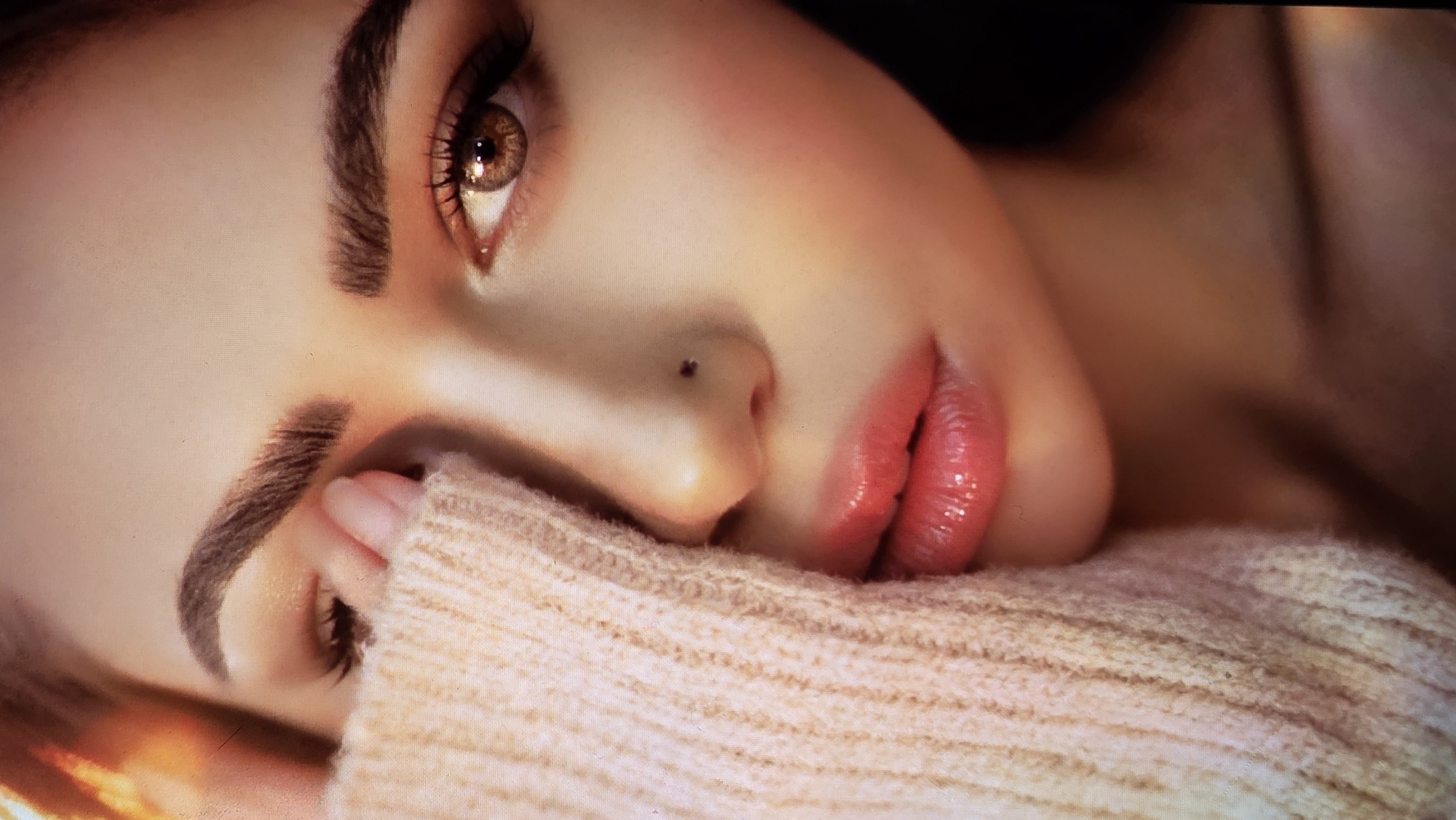 A close-up image depicts a woman lying down, with her face partially obscured by a light-colored knitted sweater. Her left eye is open and brown, with visible eyelashes and eyebrow. Her lips are red and glossy. A mole is visible on her nose. The background is dark and indistinct. There is no indication of the city of London, United Kingdom.