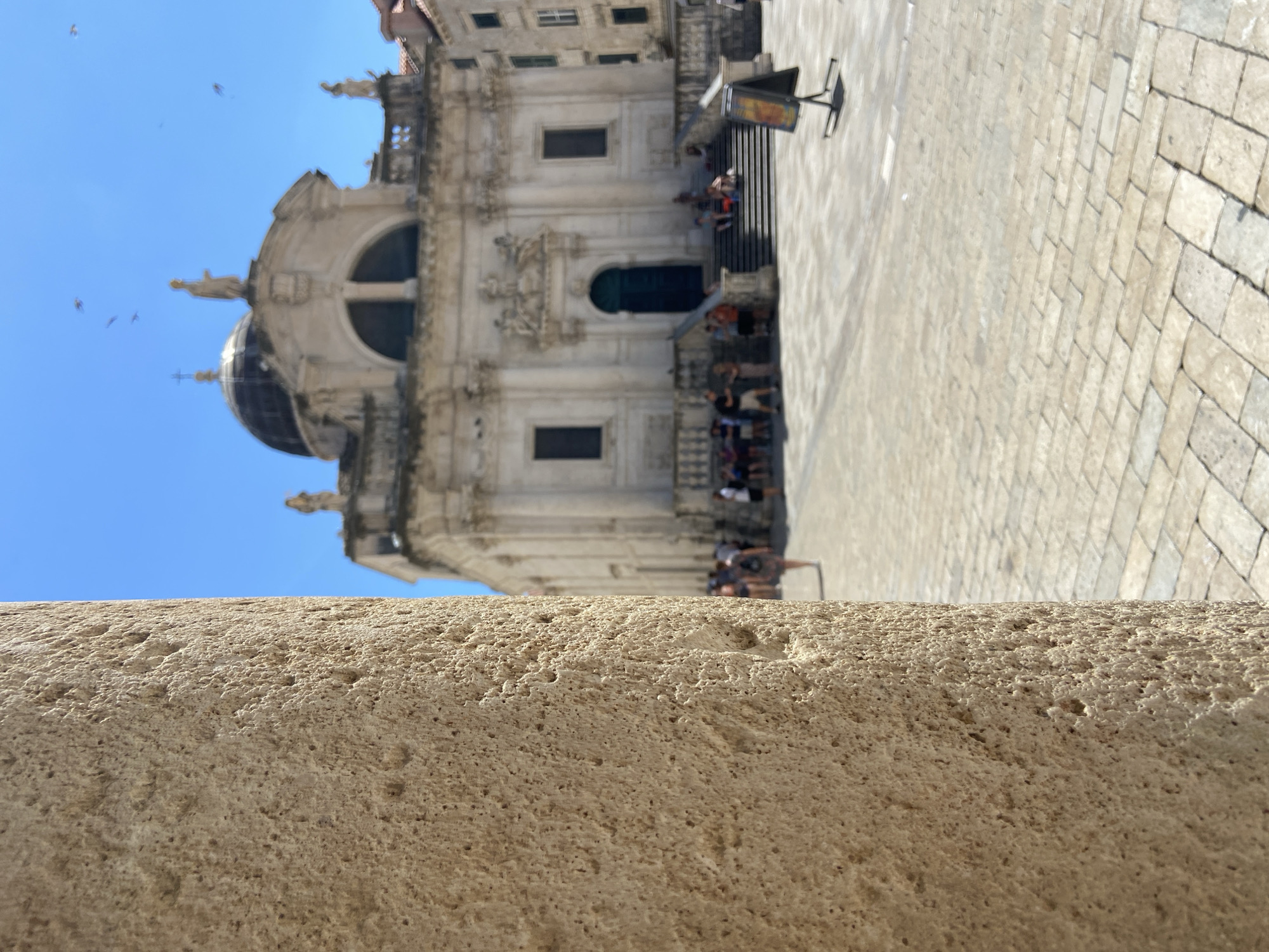 The image shows a close-up of a textured stone pillar in the foreground, obscuring the left half of the frame. In the background, a blurred scene unfolds in a city square under a bright blue sky. A domed building, likely a church with classical architectural elements, stands prominently. Several people are visible in the square, some walking, others sitting on the steps leading up to the building. A small easel with a sign is positioned to the right of the steps. The ground is paved with cobblestones. The overall scene suggests a sunny day in an historic European city, with Dubrovnik, Croatia being a strong possibility given the architectural style.