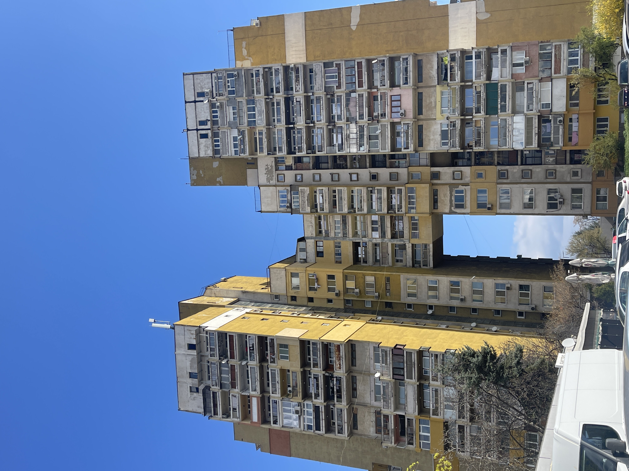 The image shows a pair of brutalist apartment buildings in Sofia, Bulgaria. The buildings are yellow and gray, with many windows. The sky is blue. There are some trees and vehicles in the lower right of the image.