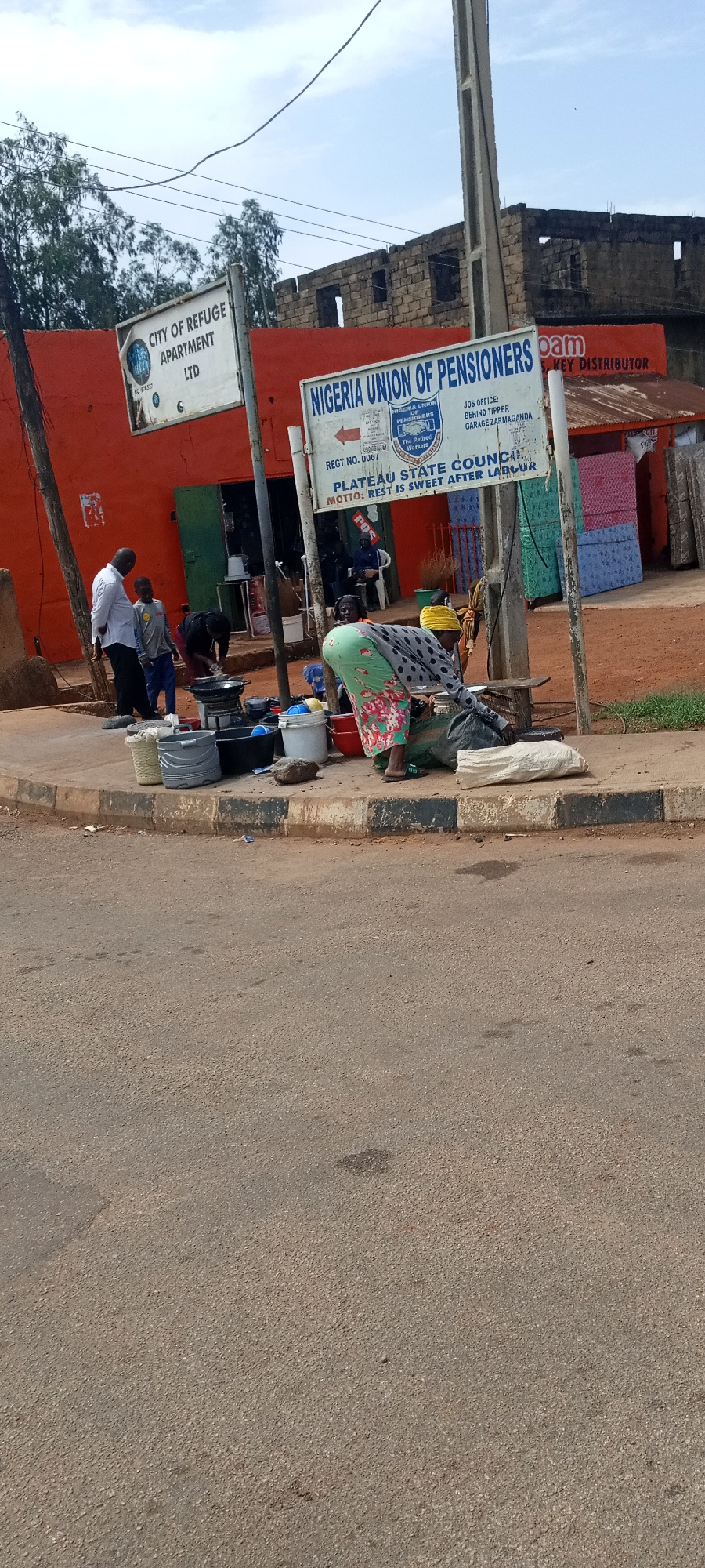 A daytime street scene in Unknown, Unknown depicts a vibrant corner with several points of interest. In the foreground, a woman in a green and floral dress bends over, engaging with items on the ground near a white sack. Behind her, a curb divides the street from a sidewalk.
On the sidewalk, there's a collection of buckets and basins, suggesting some kind of washing or cleaning activity. Further back, a group of people are gathered near a doorway of an orange building labeled "CITY OF REFUGE APARTMENT LTD". Two men, one in a white shirt and the other in a grey shirt, stand to the left, seemingly observing the scene. Near the doorway, another individual is bent over, possibly engaged in cleaning or cooking with a small cooking stove.
To the right of the orange building, there's a white sign that reads "NIGERIA UNION OF PENSIONERS," providing information about the organization's Jos office and its motto. Beyond the sign, there's a structure displaying mattresses, labeled "KEY DISTRIBUTOR."
The background features a building under construction, adding to the urban environment of the scene. A utility pole stands tall, and trees are visible in the background, providing a sense of place and context.