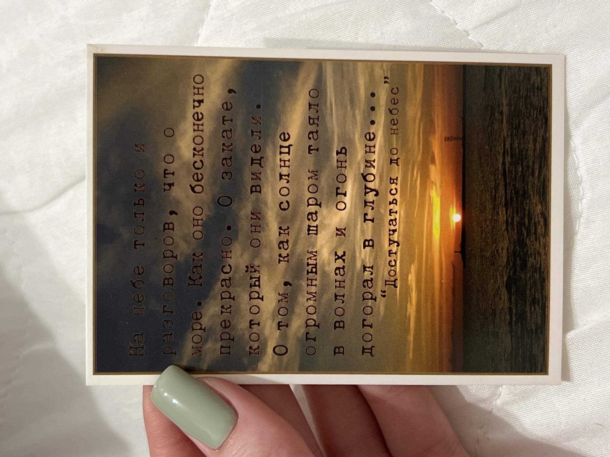 A postcard with a sunset over the ocean is held by a person's hand. The postcard has text written in Russian that says: "На небе только и разговоров, что о море. Как оно бесконечно прекрасно. О закате, который они видели. О том, как солнце огромным шаром таяло в волнах и огонь догорал в глубине... "Дотстучаться до небес"". This translates to: "All they talk about in the sky is the sea. How infinitely beautiful it is. About the sunset they saw. About how the sun, a giant ball, melted into the waves and the fire burned in the depths... 'Reaching the heavens'". The postcard is positioned on a white, textured surface, possibly a tablecloth or bedspread. The background shows the sunset with a lighthouse in the distance. No specific location in Kazan, Russia, can be identified from the image.
