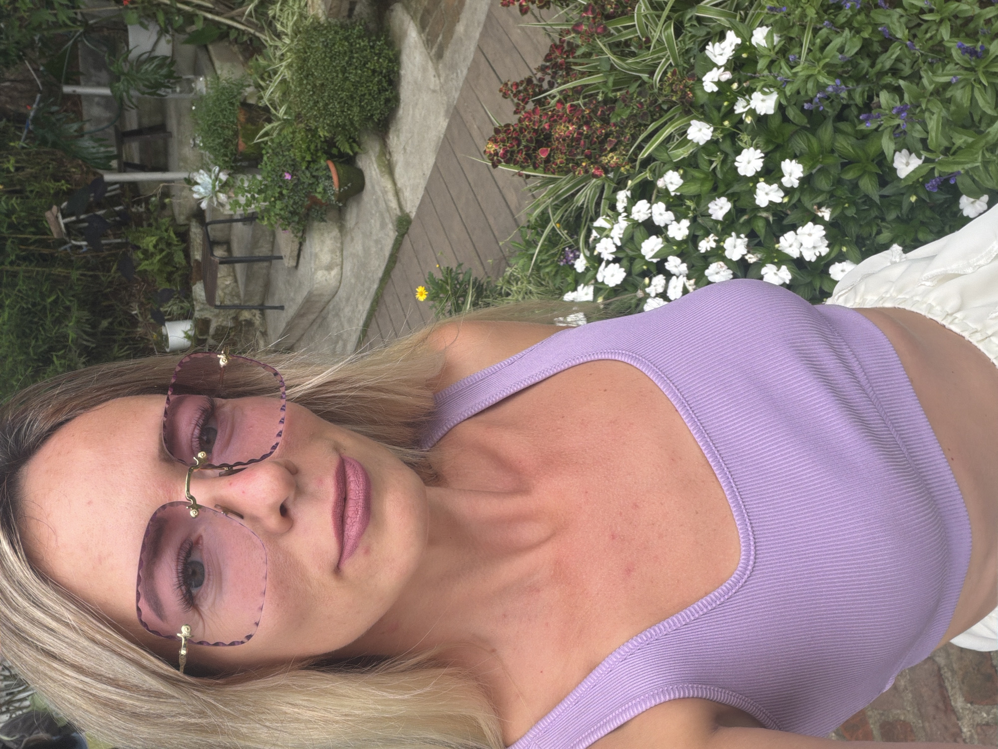The media file shows a close-up, selfie-style shot of a blonde woman with fair skin, wearing large, rose-tinted sunglasses and a lavender ribbed tank top. Her lips are painted a matte pink. The background suggests an outdoor setting with lush greenery and white flowers, possibly a garden. In the background, there are also glimpses of stone steps, wooden decking, and potted plants. The lighting indicates it is daytime. There is no visible text. The overall impression is a casual, relaxed moment captured outdoors.