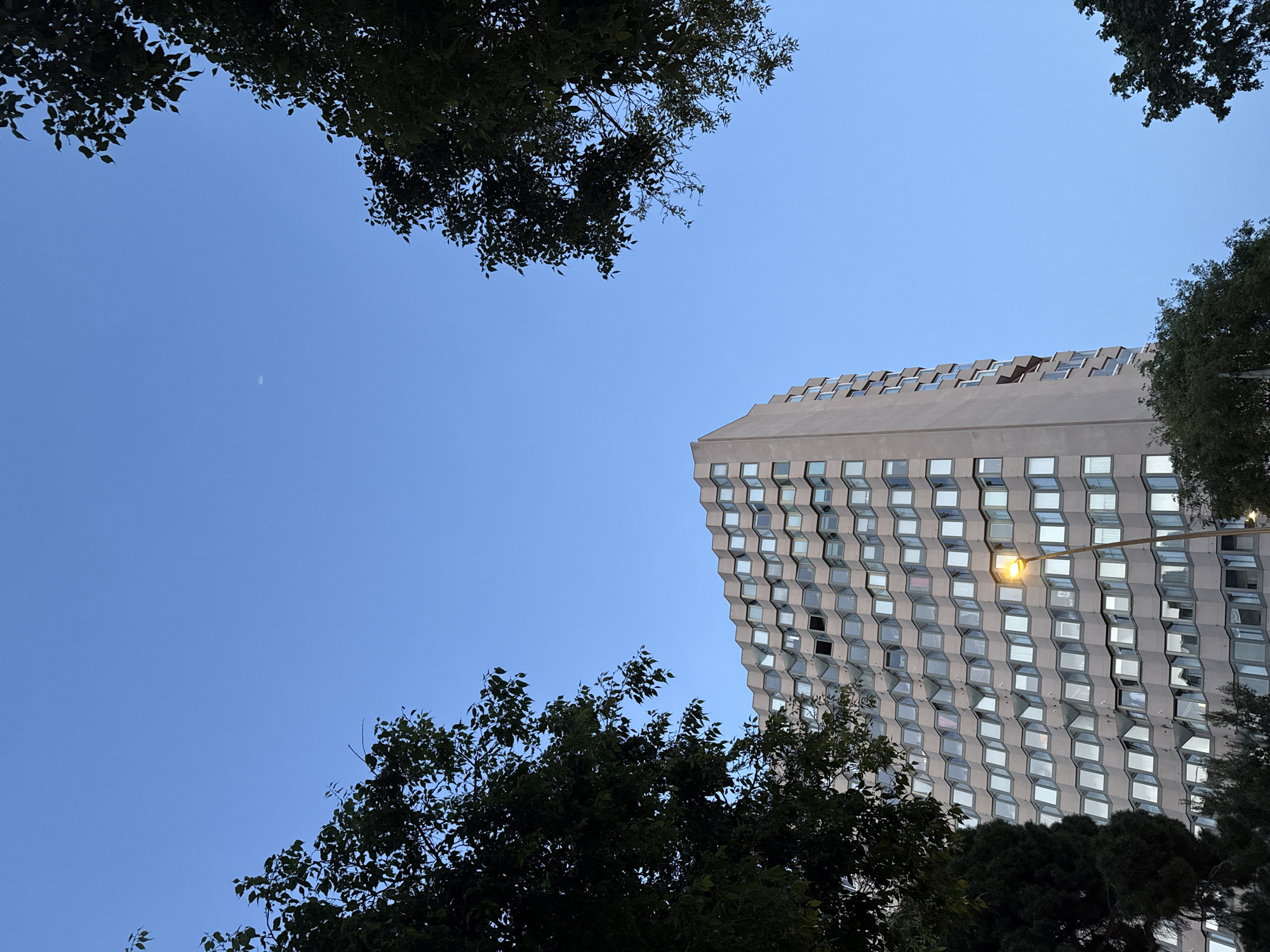 The image shows a view upwards towards the sky in Barcelona, Spain. Tree foliage appears in the top left and bottom of the frame. A large, modern-looking building with multiple windows is visible on the right. A streetlamp is attached to the building. In the upper left, the moon is visible in the clear blue sky.