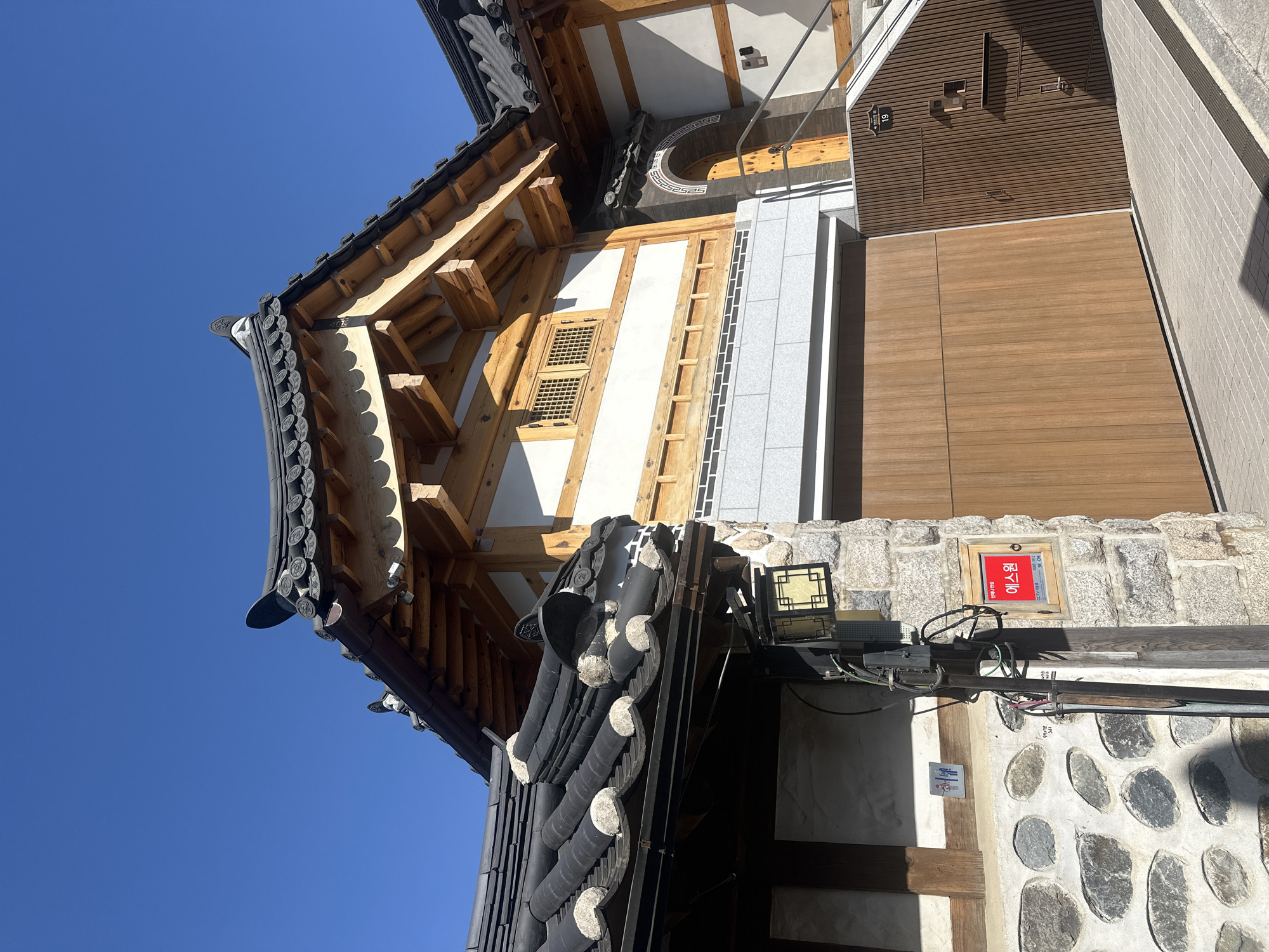 The image captures a traditional Korean building in Seoul, South Korea, characterized by its distinct tiled roof with rounded edges, intricate wooden beam construction, and white-painted walls. The architecture is a blend of historical aesthetics with modern materials like the light-colored stone foundation and sleek wooden paneling. A red rectangular sign, possibly a business or address marker, is visible on the stone wall. Utility elements like electrical wires and a meter box are also present, juxtaposing the old-world charm with modern infrastructure. The clear blue sky forms the backdrop. The building appears to be a renovated or newly constructed structure blending traditional Korean architectural elements with contemporary designs.