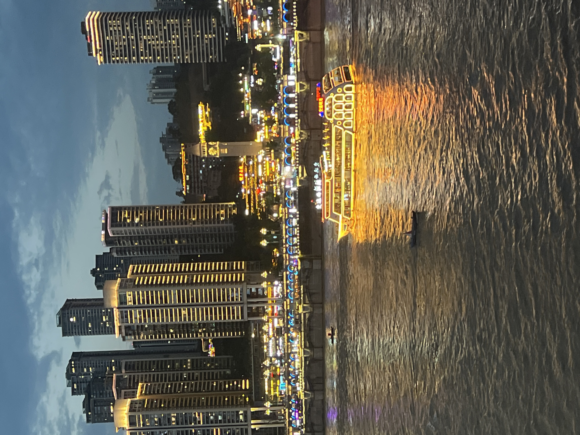 The image captures a twilight scene along a waterfront in Nan'an District, China, likely Chongqing. A large, illuminated tour boat, adorned with yellow and red lights, cruises on a wide, rippling body of water. The boat has multiple decks and appears to be carrying passengers. In the background, a densely populated cityscape rises, featuring numerous tall, modern buildings with many windows lit up in yellow and white. A pedestrian bridge, also illuminated with colorful lights, spans the water in the middle ground. Smaller boats, one with two occupants, are also visible on the water. The sky above is a dusky blue with scattered clouds. The overall impression is one of a vibrant, urban waterfront at dusk.