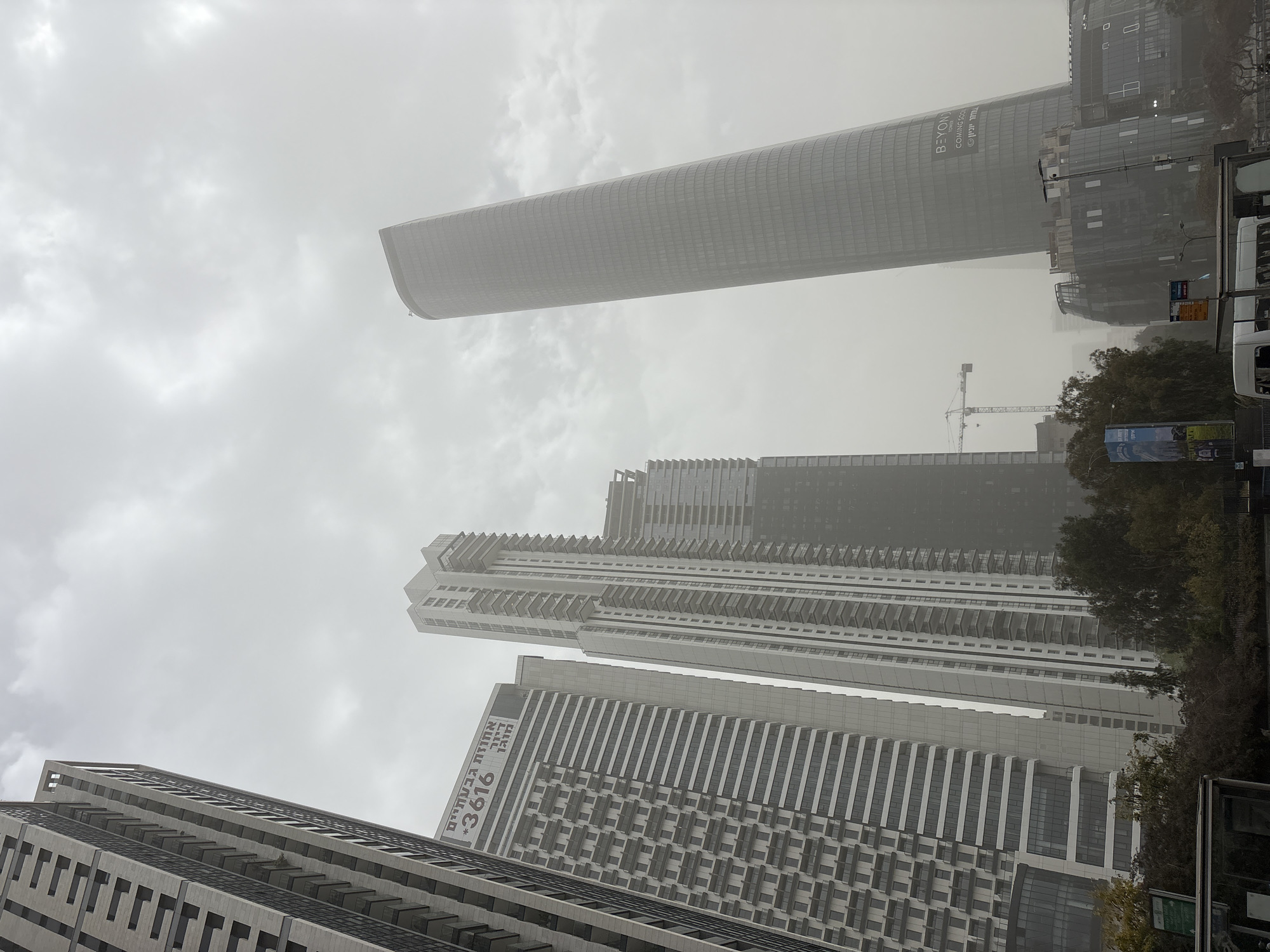 The image displays a dense urban landscape in Ramat Gan, Israel, under a heavily overcast and hazy sky. The low-angle perspective looks up at multiple towering modern buildings, creating an impression of scale and ongoing development.
A prominent feature is a tall, cylindrical glass skyscraper in the background, partially obscured by the haze. Visible on its facade are the English words "BEYOND COMING SOON" and Hebrew text "ביונד פארק אריאל שרון" (Beyond Ariel Sharon Park). In the mid-ground and foreground, several other high-rise buildings are visible, characterized by repetitive patterns of windows and balconies. One building prominently displays Hebrew text "אחוזות גבעתיים *3616" (Achuzot Givatayim *3616).
A construction crane can be seen in the distance, indicating active building projects. Trees are visible on the right side of the image, breaking the urban monotony, and parts of vehicles, possibly buses, are partially visible on the far right. The cloudy, muted lighting suggests daytime but not bright sunshine, contributing to a somewhat somber or industrial atmosphere. No specific human activities or emotions are discernible; the scene is purely architectural and environmental.