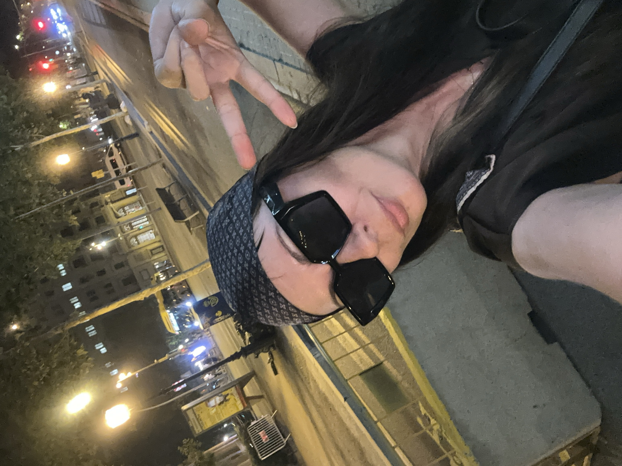 In a street-level, night-time photograph taken in Barcelona, Spain, a woman poses for a selfie. She is wearing large, black, square-framed sunglasses and a patterned bandana on her head. Her right hand is raised, displaying a peace sign. She is looking at the camera, her mouth slightly open. She is wearing a dark top.
The background shows a street scene at night. Streetlights illuminate the scene, casting a warm glow. Buildings with illuminated windows are visible in the distance. A bus is stopped at what appears to be a bus stop, and there are other vehicles and pedestrians in the background. The street has a marked crosswalk, and a bench is visible on the sidewalk. A metal barrier is also present. The overall impression is of a busy urban environment at night.